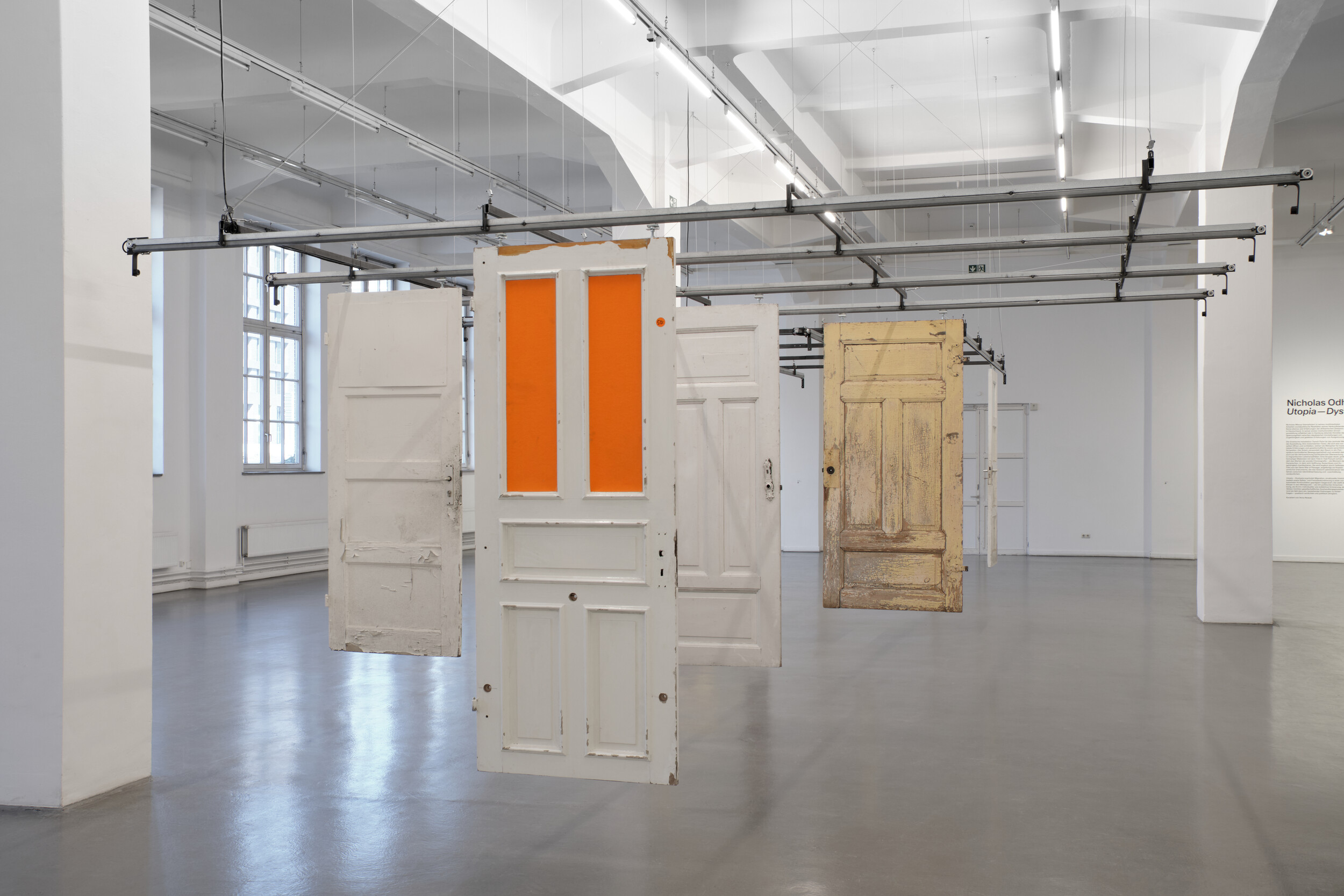 In a bright exhibition space with large windows, several old, differently styled doors hang freely in the room from a metal structure. Some doors feature colored or textured glass panels, while others show signs of wear and peeling paint.