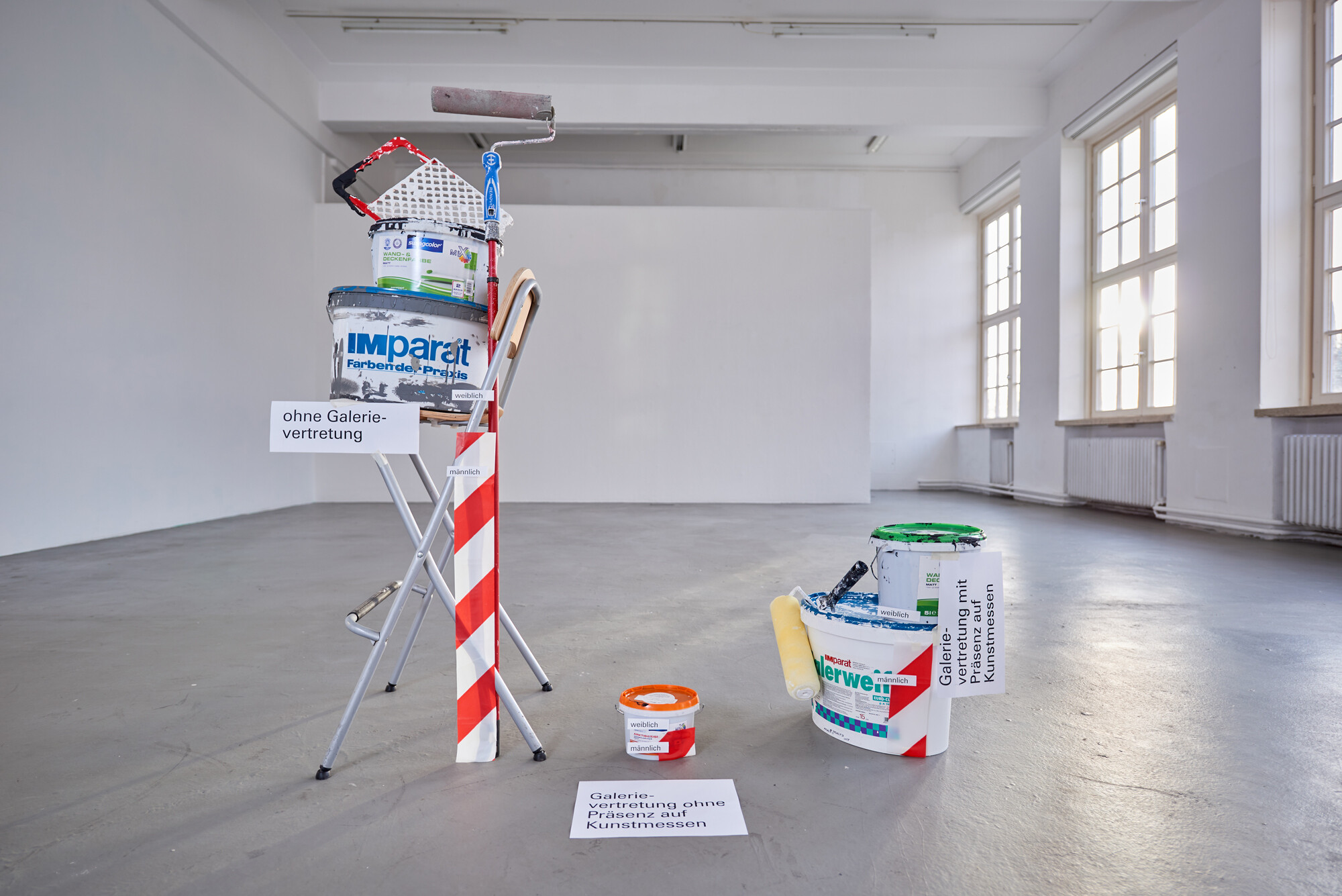 Installation in an empty, bright exhibition room: Paint buckets, rollers, and tools are arranged on a ladder and on the floor, some labeled with signs. Daylight enters through large windows.