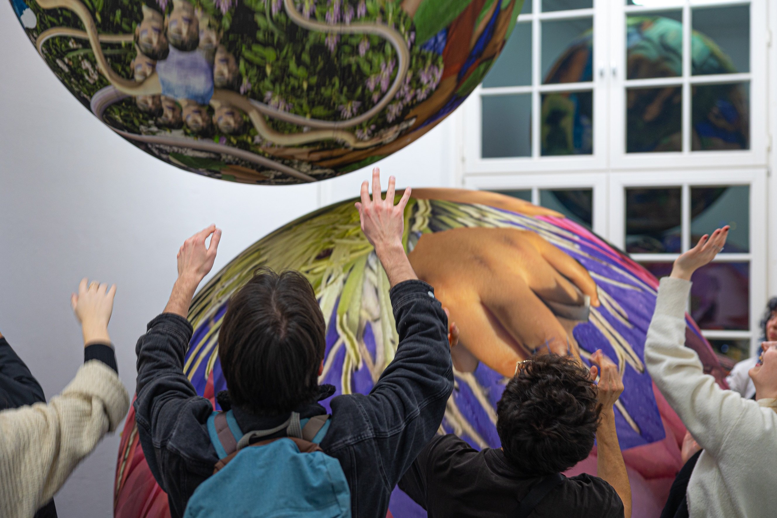 Several people stand in an exhibition space with white walls, raising their arms toward two large suspended spherical objects. The spheres are printed with colorful, collage-like imagery, including plants, figures, and body details.