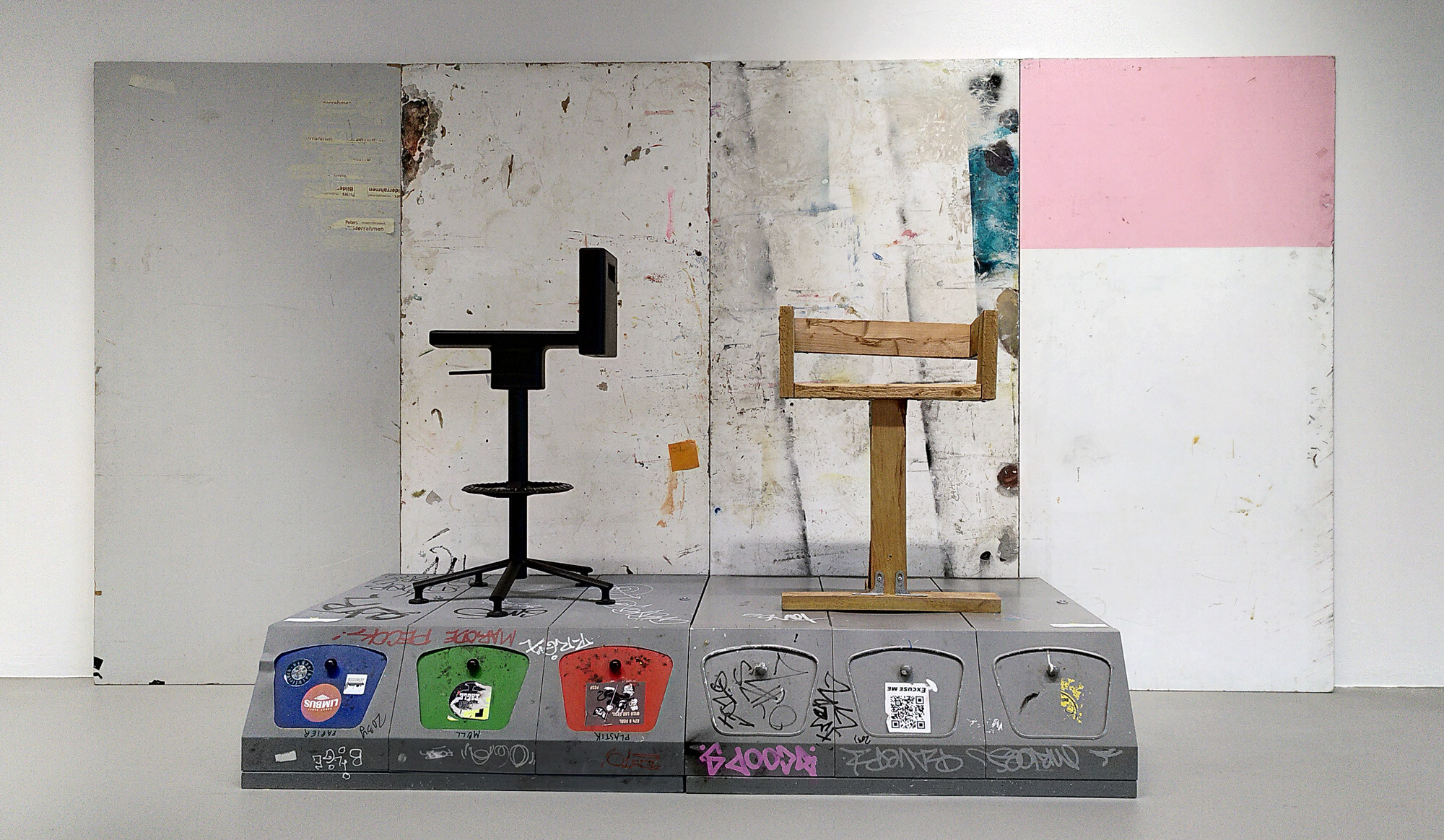 Two different stools stand on a gray pedestal covered with graffiti and stickers. The left stool is black and modern with a swivel base, while the right one is made of light wood with a simple, angular design. Behind them is a multi-panel wall surface, partly painted and marked.