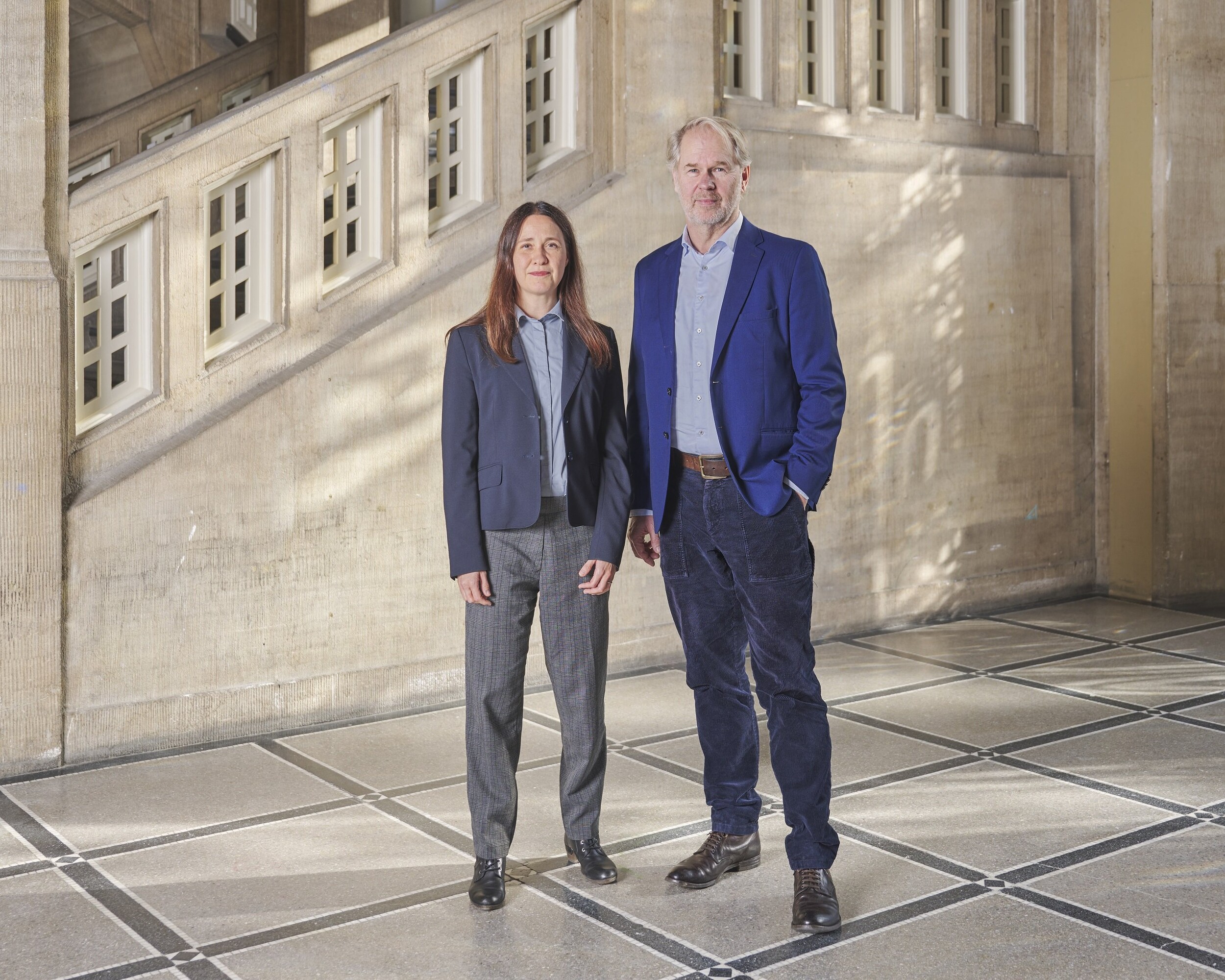 Two adults stand side by side in a bright, historic interior with stone walls and a geometric floor pattern, facing the camera.