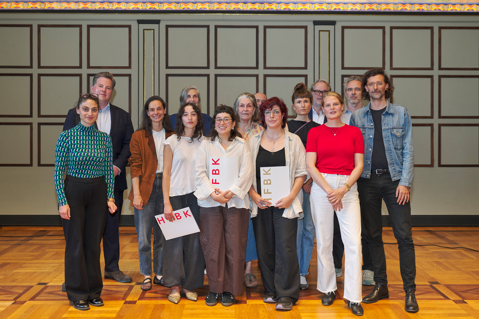 Group of around fifteen people standing together in a historic hall with patterned wall panels and wooden floor, some holding white boards with red lettering.