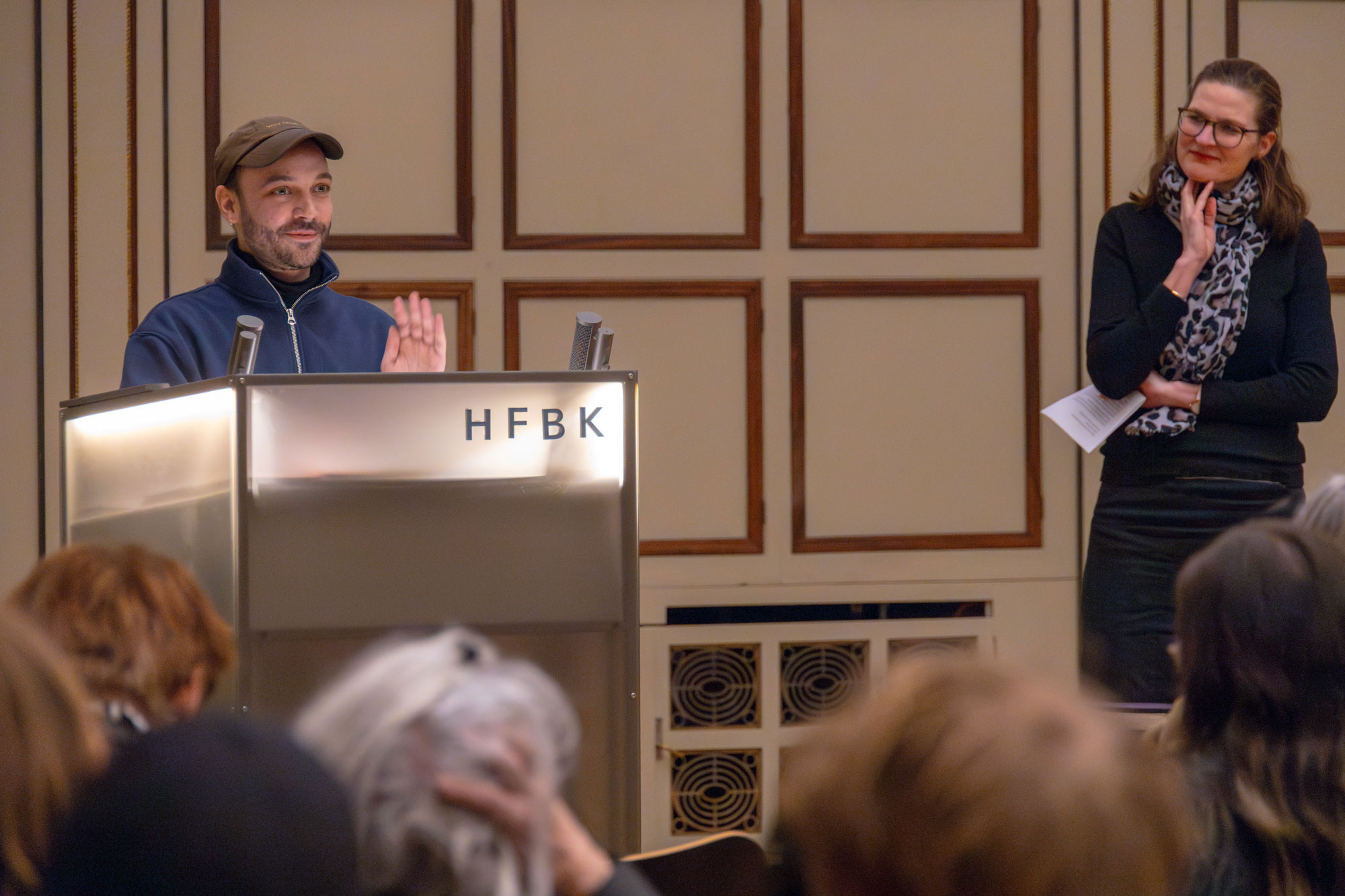 A person speaks at a lit lectern labeled “HFBK” in front of an audience; another person stands to the side holding notes, in a formal interior setting.