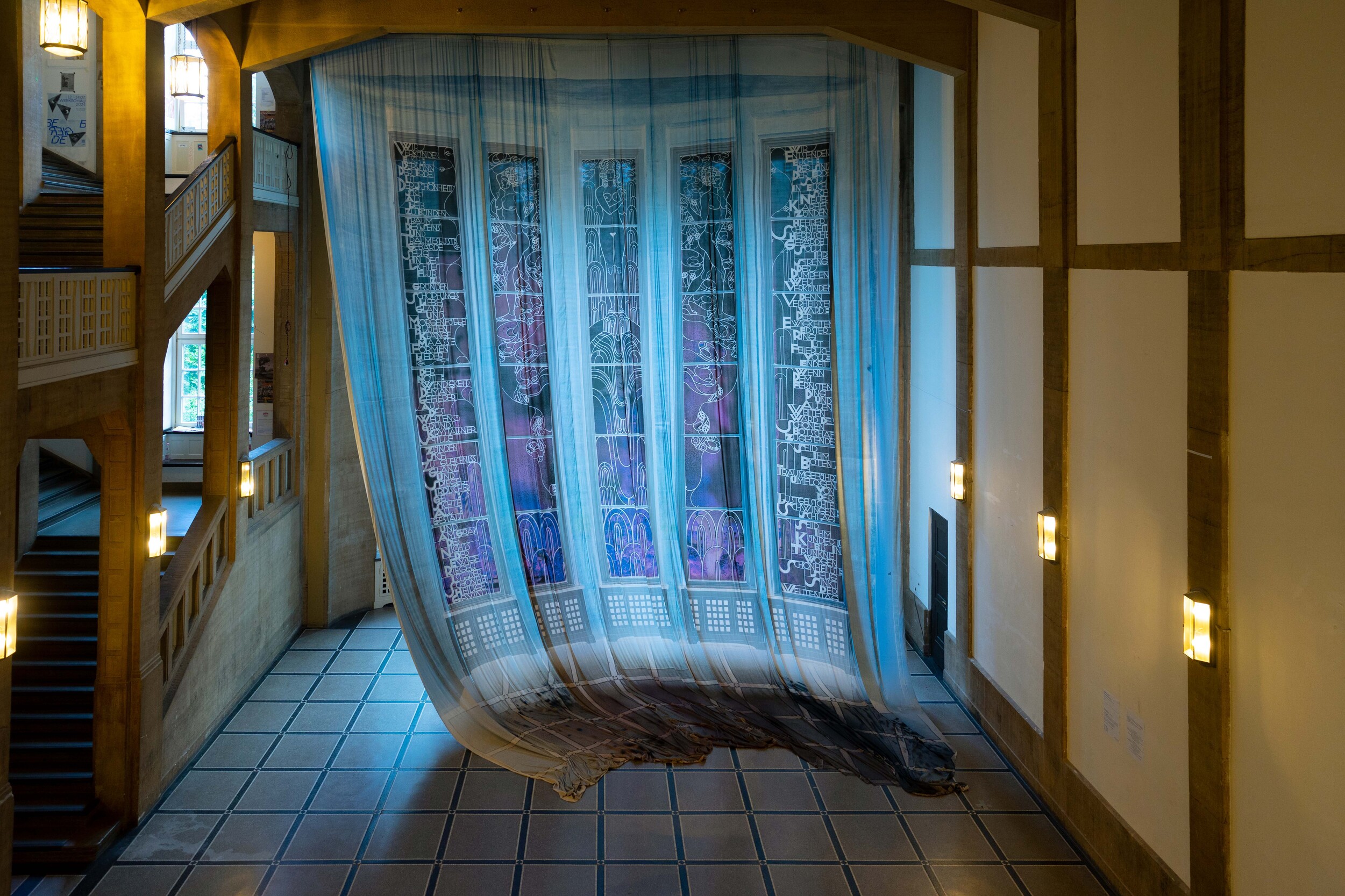 Interior view of a tall, wood-paneled stair hall with side staircases and warm wall lights. In front of a large multi-pane window hangs a semi-transparent bluish fabric curtain that drapes down to the tiled floor, softly filtering the light.