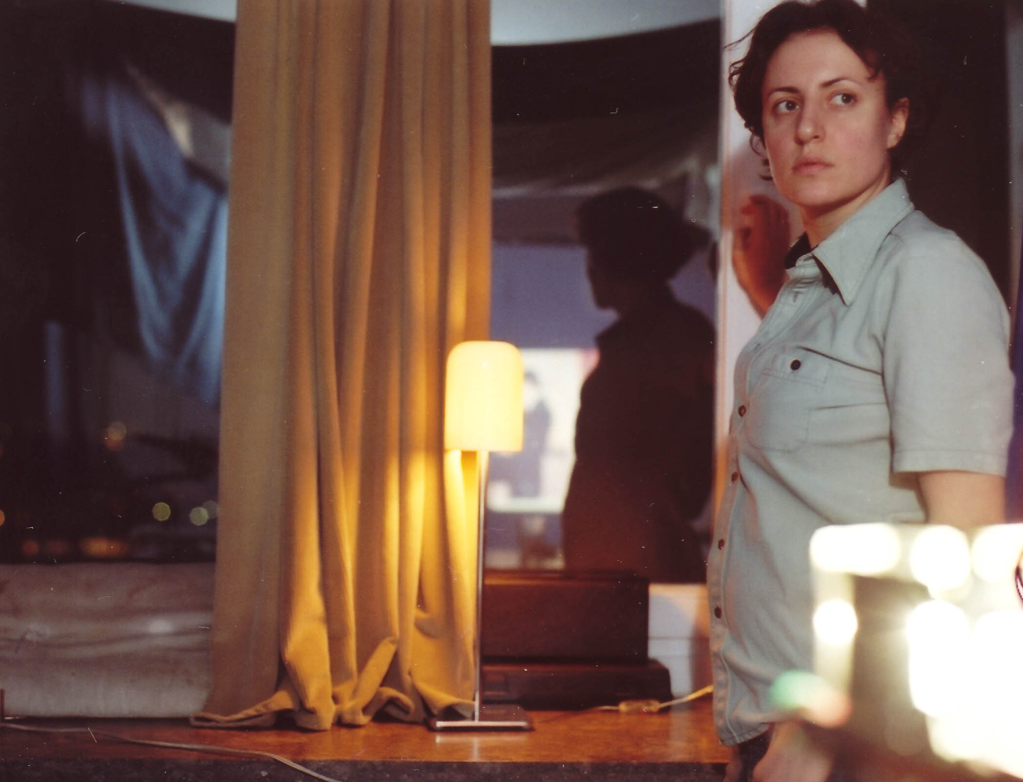 Interior space with a curtain, table lamp, and mirror; a person stands in profile in the foreground, with their reflection visible in the background, illuminated by warm light.