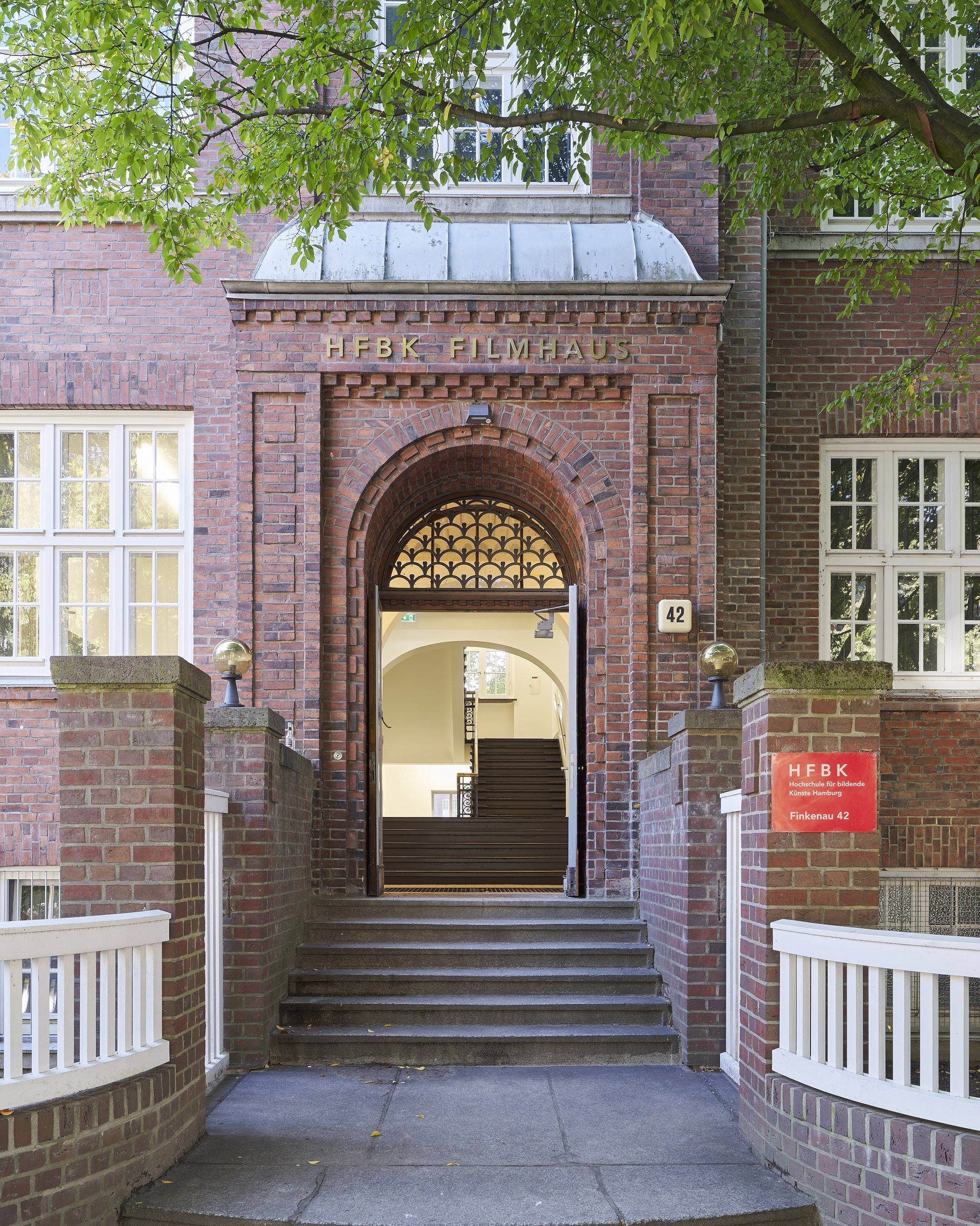 Brick building with a central entrance portal labeled “HFBK Filmhaus”; a staircase leads through the open archway into the interior, surrounded by trees.