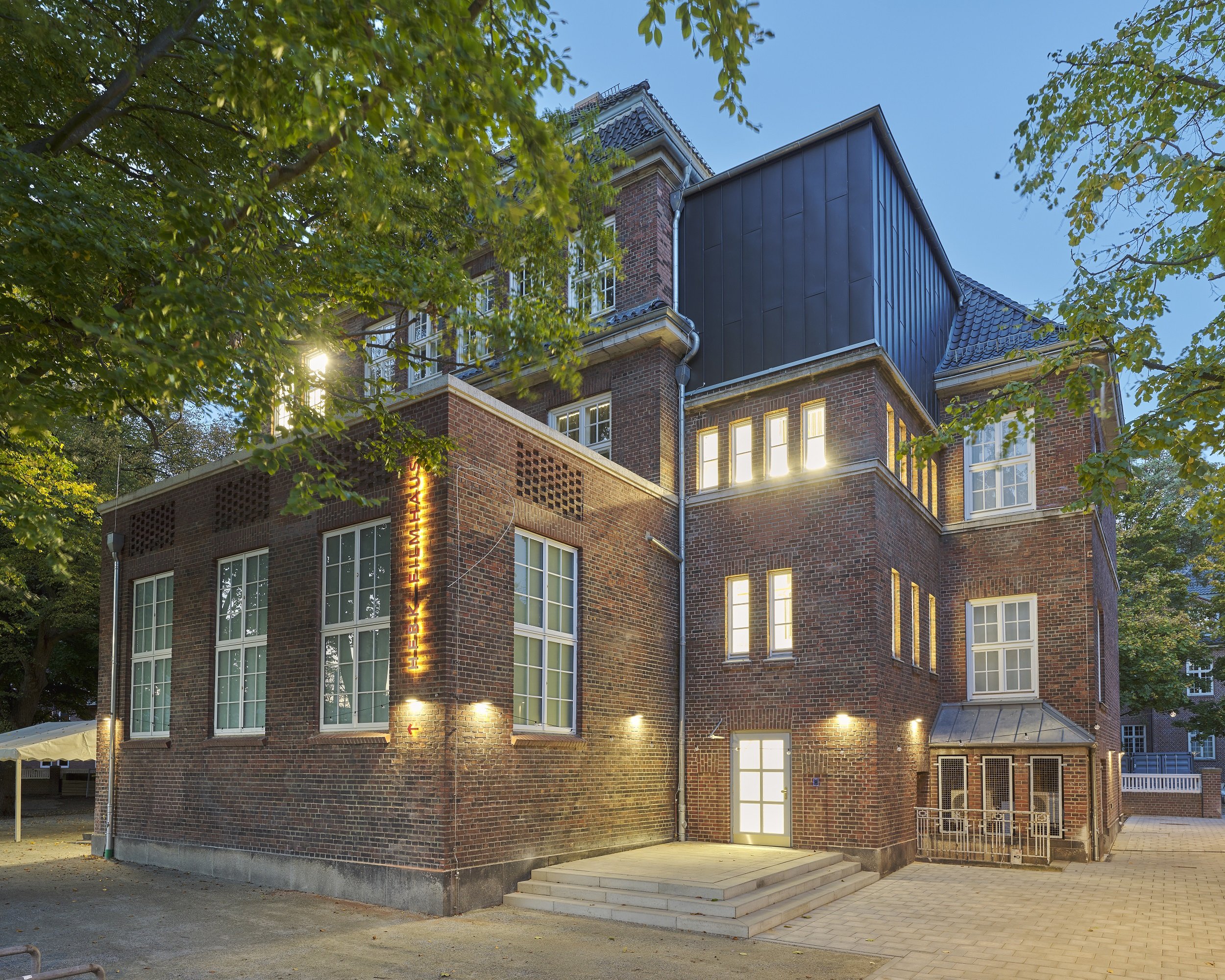 Exterior view of a multi-story brick building with illuminated windows and a modern upper extension; trees frame the building in the foreground.