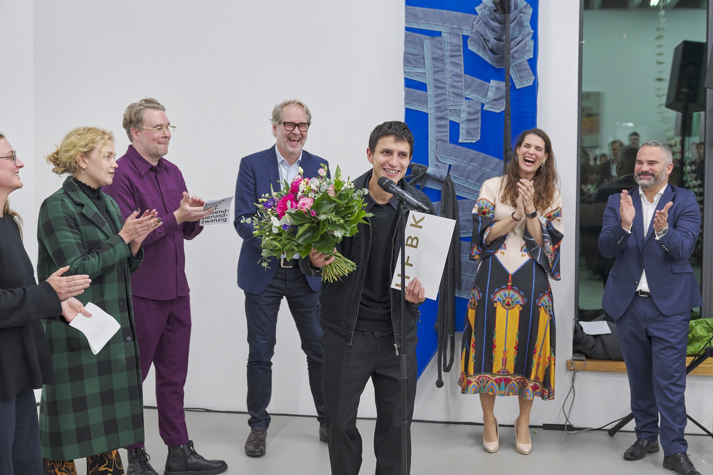Eine Person spricht an einem Mikrofon und hält einen Blumenstrauß; mehrere Personen stehen daneben und applaudieren. Im Hintergrund ist ein textiles, blaues Wandobjekt zu sehen.