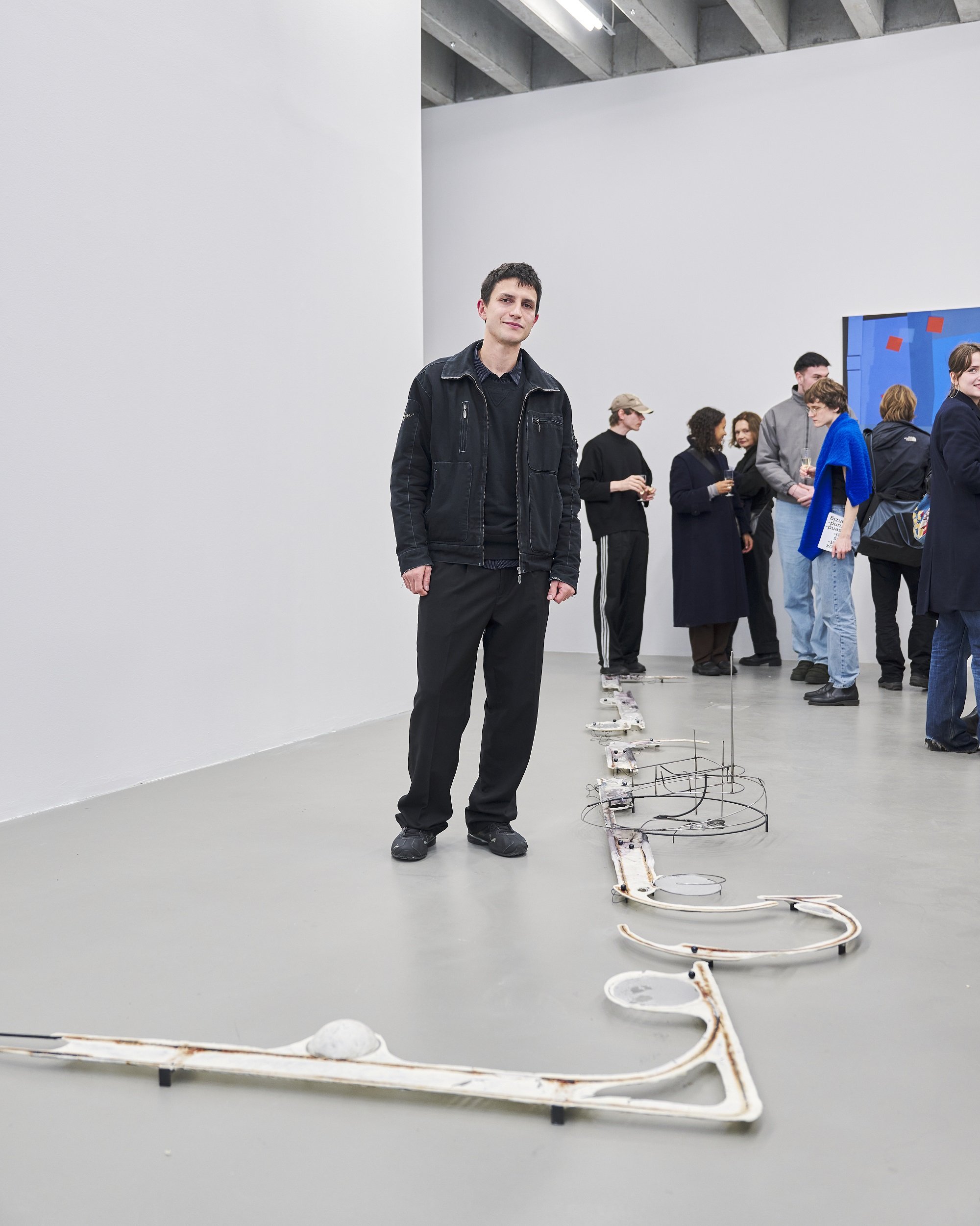 Eine Person steht in einem hellen Ausstellungsraum nahe einer langgezogenen Skulptur am Boden; im Hintergrund unterhalten sich mehrere Besucherinnen und Besucher vor einer Wandarbeit.