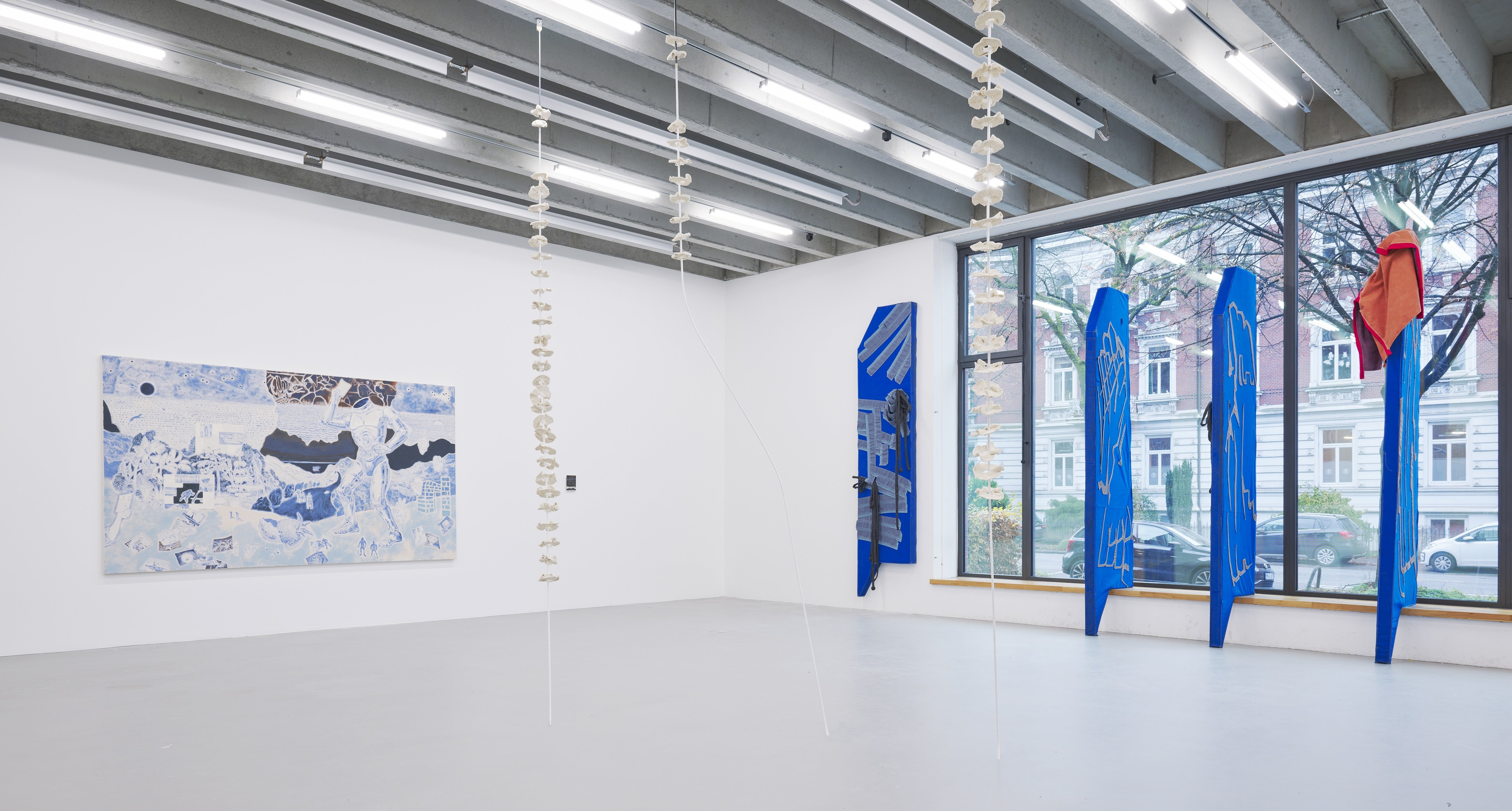 Large, bright exhibition space featuring multiple artworks: blue textile sculptures placed near the windows, a wall-mounted drawing on the left, and two light-colored objects hanging vertically from the ceiling.