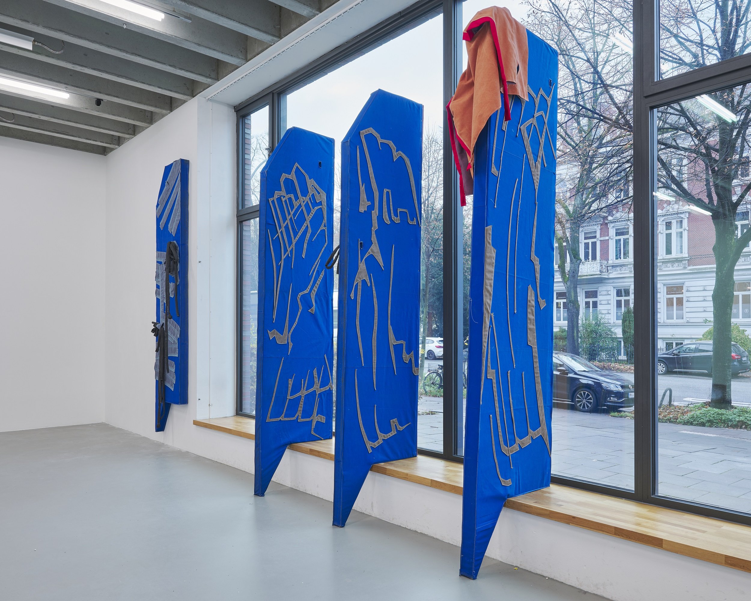 Installation of several tall blue textile sculptures with stitched line patterns, standing upright in front of large windows inside an exhibition space; one sculpture is draped with red fabric at the top.