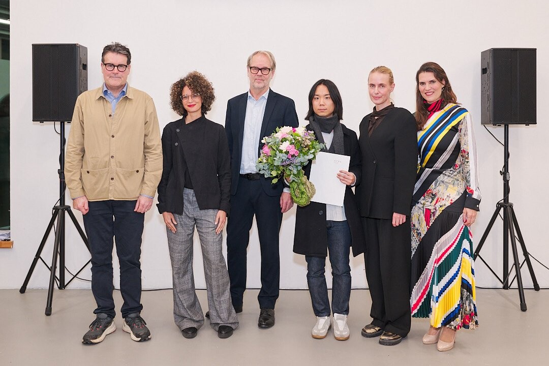 Seven people stand side by side in front of a white wall in an exhibition space, flanked by two speakers; one person is holding a bouquet of flowers and a document.