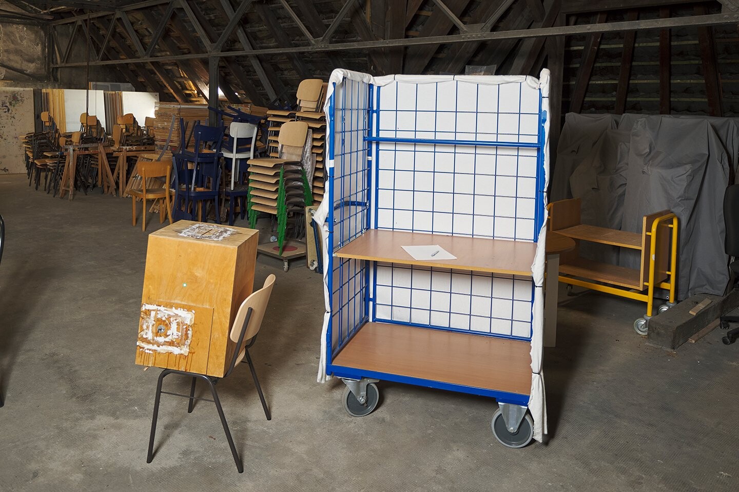 Innenraum mit gestapelten Stühlen und Möbeln; im Vordergrund stehen ein hölzerner Wahlkasten mit Sitzgelegenheit sowie ein mobiler, gitterartiger Wahlstand auf Rollen mit Ablageflächen.