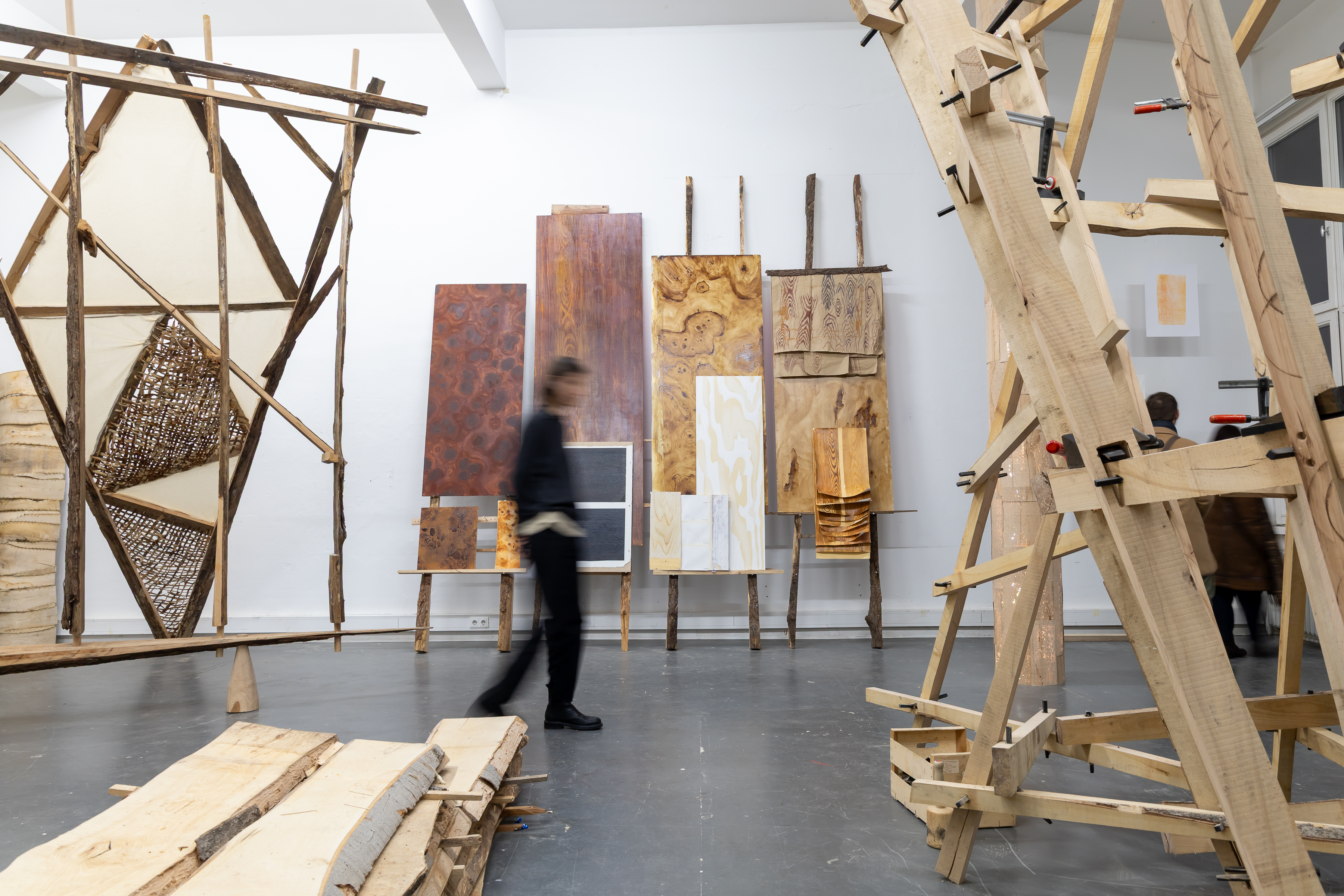 Exhibition space featuring large wooden sculptures and wall works made from different types of wood and finishes. A person walks blurred through the room, and wooden planks lie on the floor in the foreground.