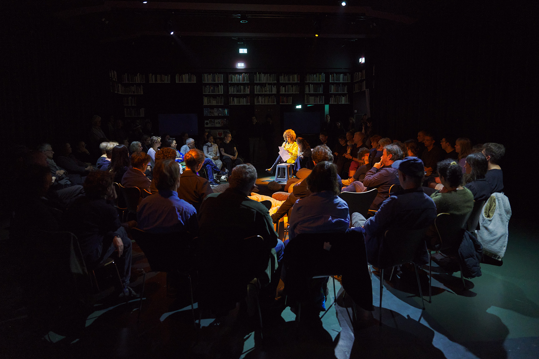 A person sits on a chair in the center of a dimly lit room, reading from a sheet of paper under a warm spotlight. The audience is seated in a circle around them, with bookshelves visible along the walls in the background.