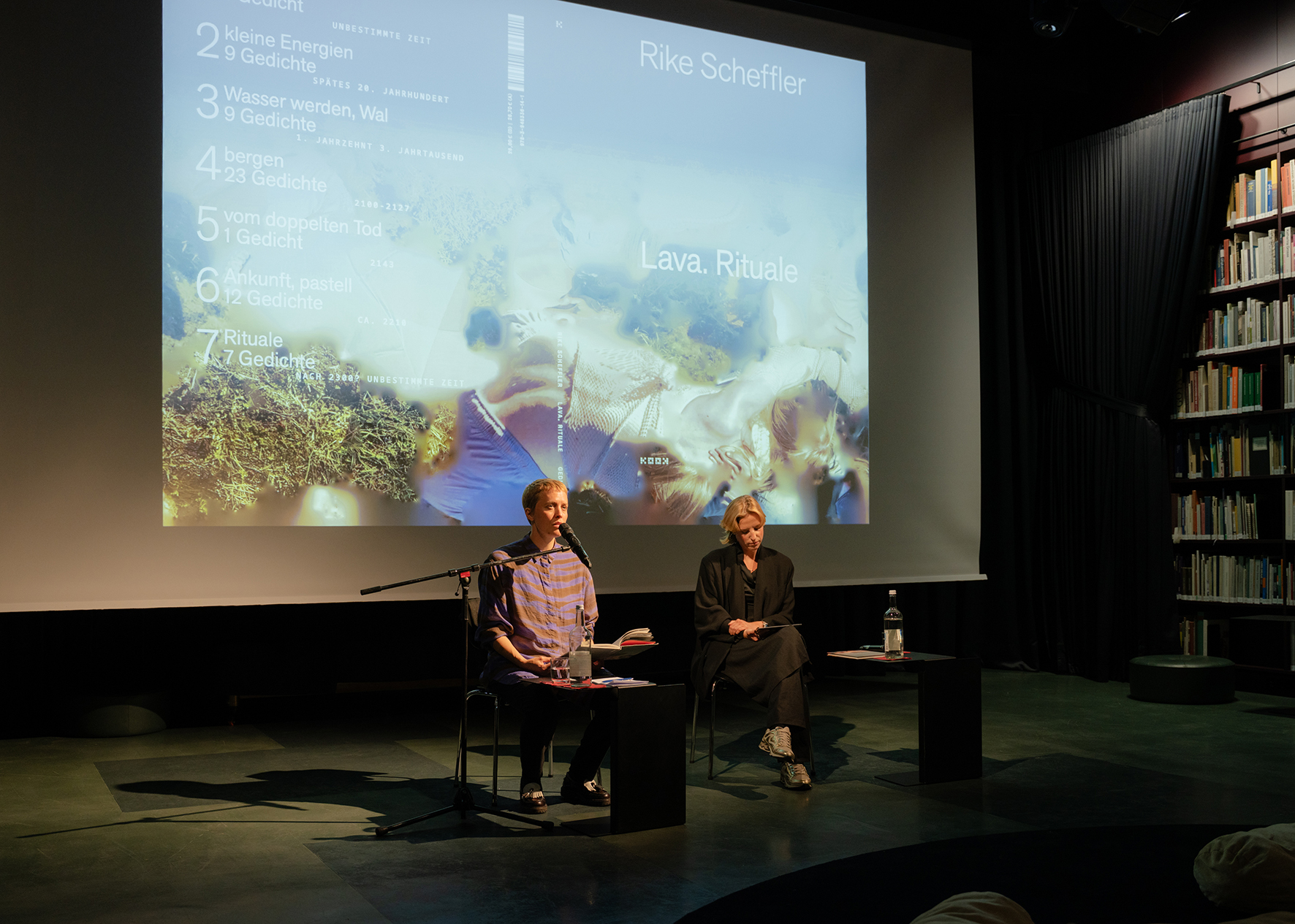Two people are seated on a stage in front of a large projection screen displaying the book cover of Lava. Rituale by Rike Scheffler. One person reads from a book into a microphone while the other listens; small tables with glasses of water stand beside them. Tall bookshelves are visible on the right side of the room.