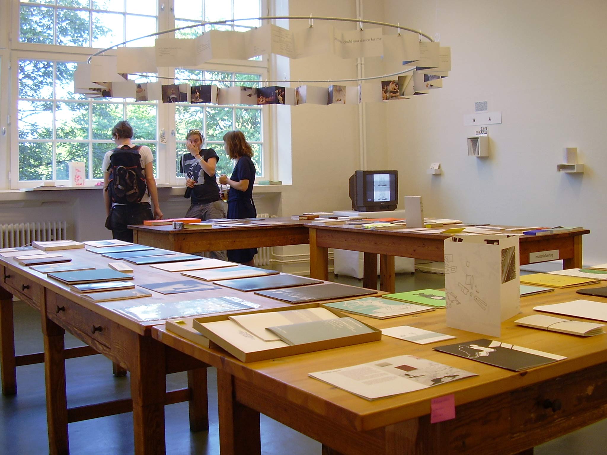 Bright exhibition or project space with large windows and several wooden tables displaying numerous books and publications. Three people stand by the window in conversation. Above the tables hangs a circular installation with suspended sheets or photographs.