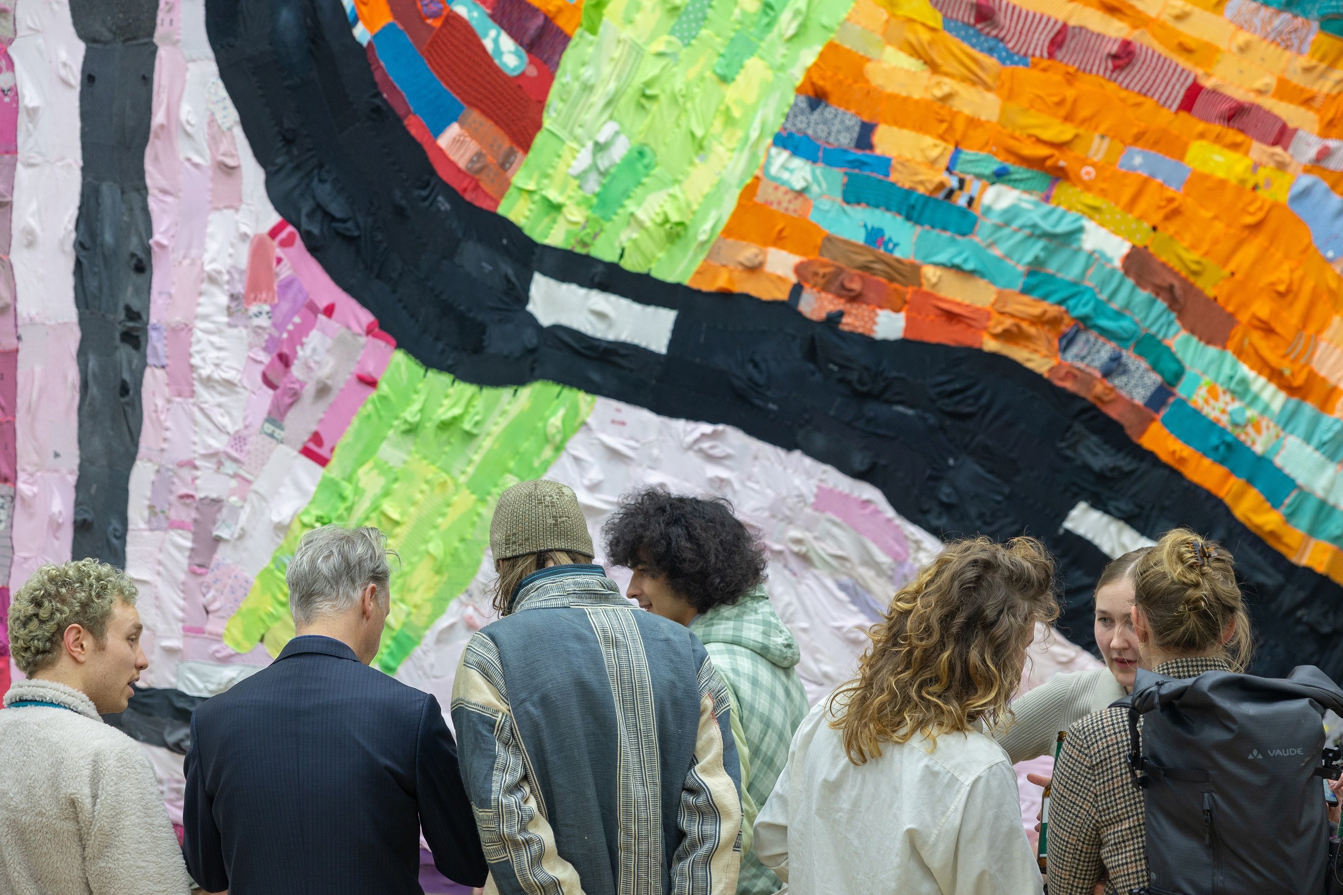 Sieben Personen stehen vor einer bunten Wand aus unterschiedlich farbigen Stoffstreifen.