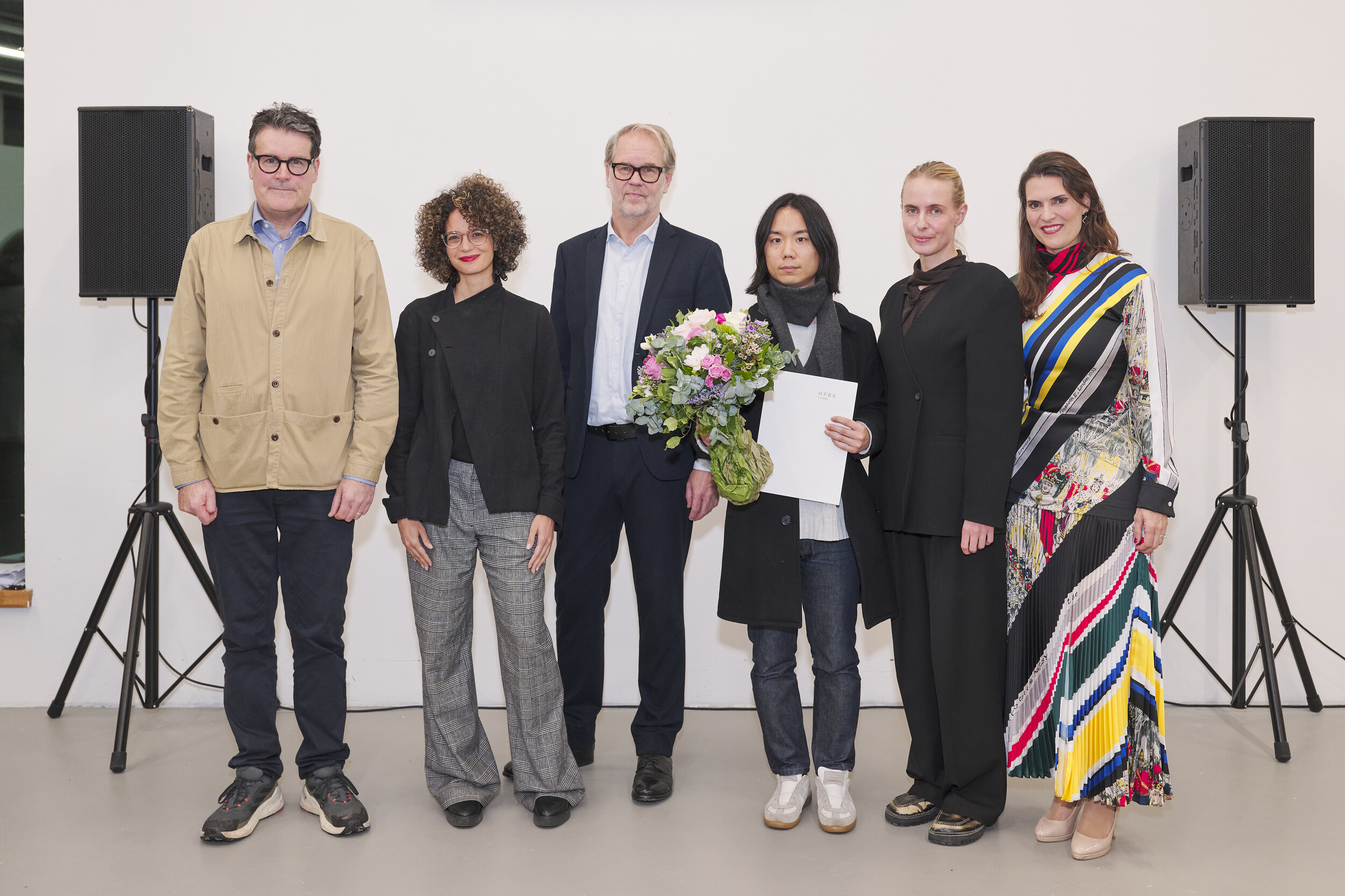 Six people stand side by side in front of a white wall between two speakers on stands. The person in the center holds a bouquet of flowers and a document. The group is dressed in formal or semi-formal attire and poses together for a photograph indoors.