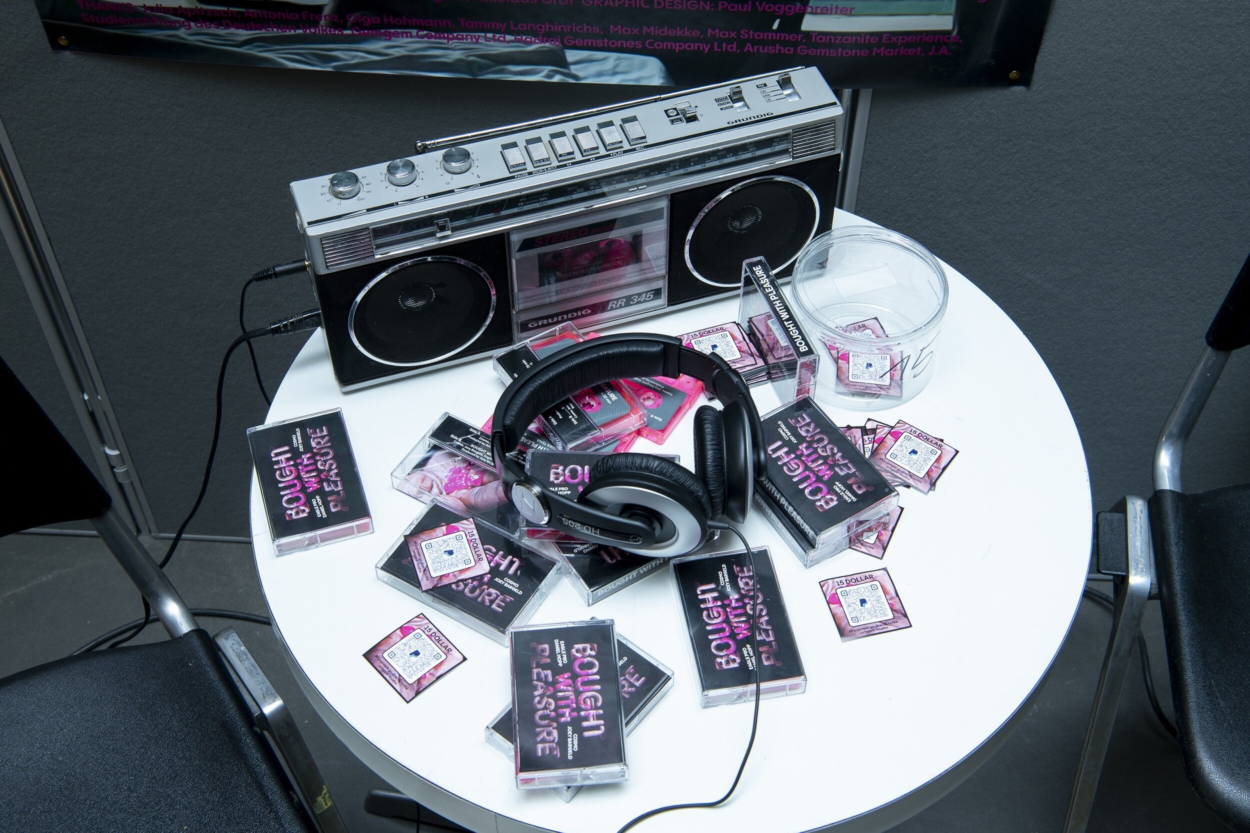A cassette recorder from the 1980s stands on a round white table. Several cassette cases with pink and white lettering on a black background lie on the table. Headphones are also on the table.