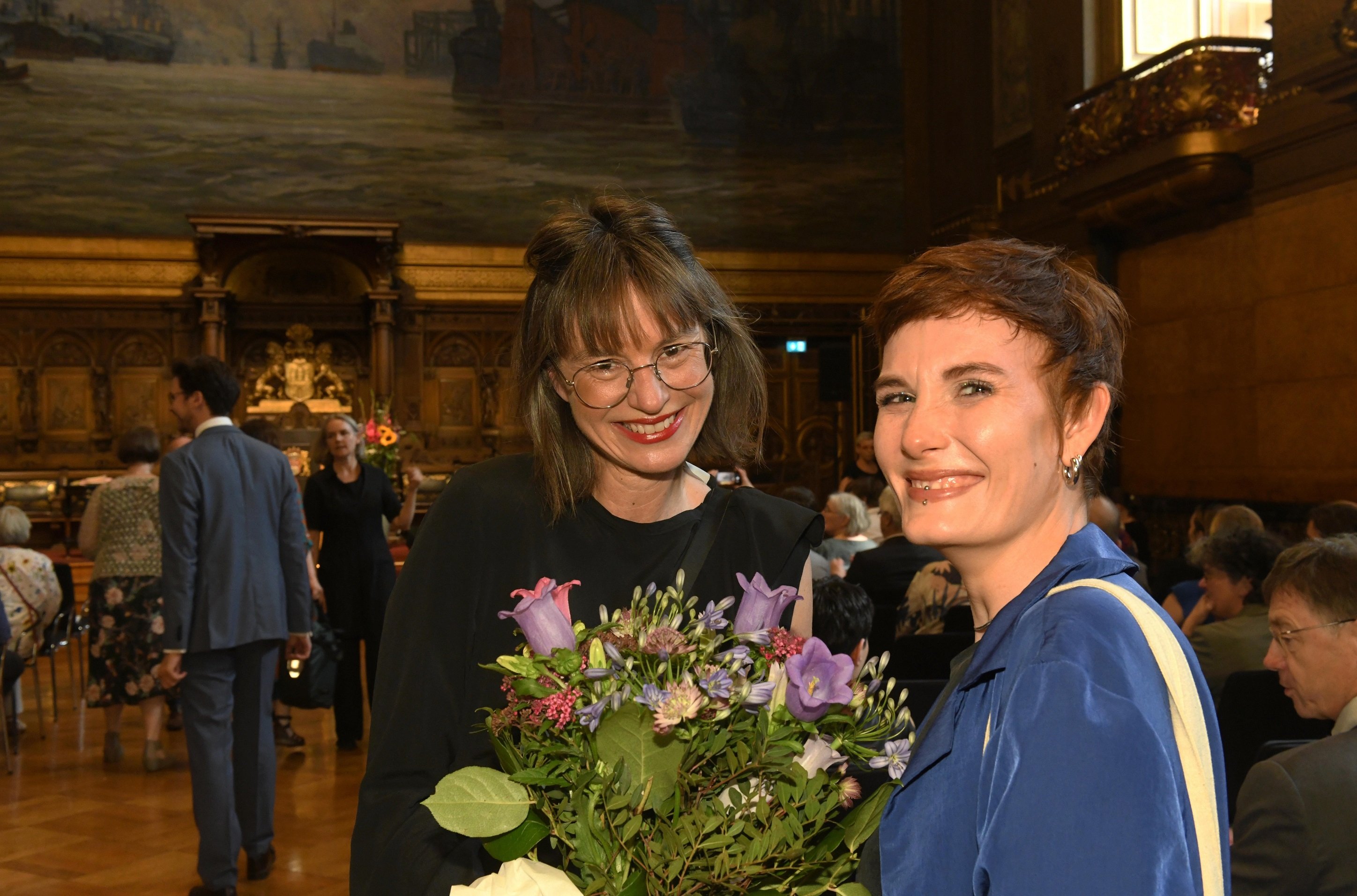 Zwei Frauen stehen in einem festlich beleuchteten historischen Saal und lächeln in die Kamera. Eine von ihnen hält einen Blumenstrauß mit violetten und rosafarbenen Blüten. Im Hintergrund sind weitere Gäste zu sehen, die auf Stühlen sitzen oder sich unterhalten; der Raum ist mit Holzvertäfelungen und einem großen Wandgemälde gestaltet.