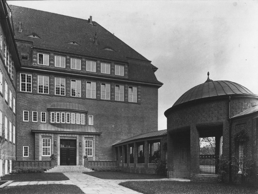 Historische Schwarzweißaufnahme eines mehrgeschossigen Backsteingebäudes mit steilem Dach und symmetrischer Fensterfront; rechts daneben ein runder Pavillon mit Kuppeldach und offener Säulenhalle, verbunden durch einen überdachten Gang.
