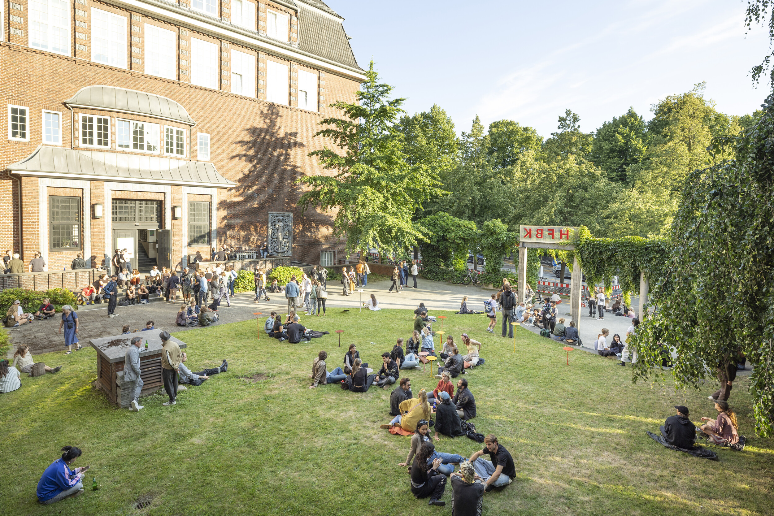 Blick auf den begrünten Innenhof eines Backsteingebäudes, in dem zahlreiche Menschen auf dem Rasen sitzen, stehen und sich unterhalten; rechts steht eine mit Efeu bewachsene Pergola mit dem Schriftzug „HFBK“, im Hintergrund Bäume und sommerliches Licht.