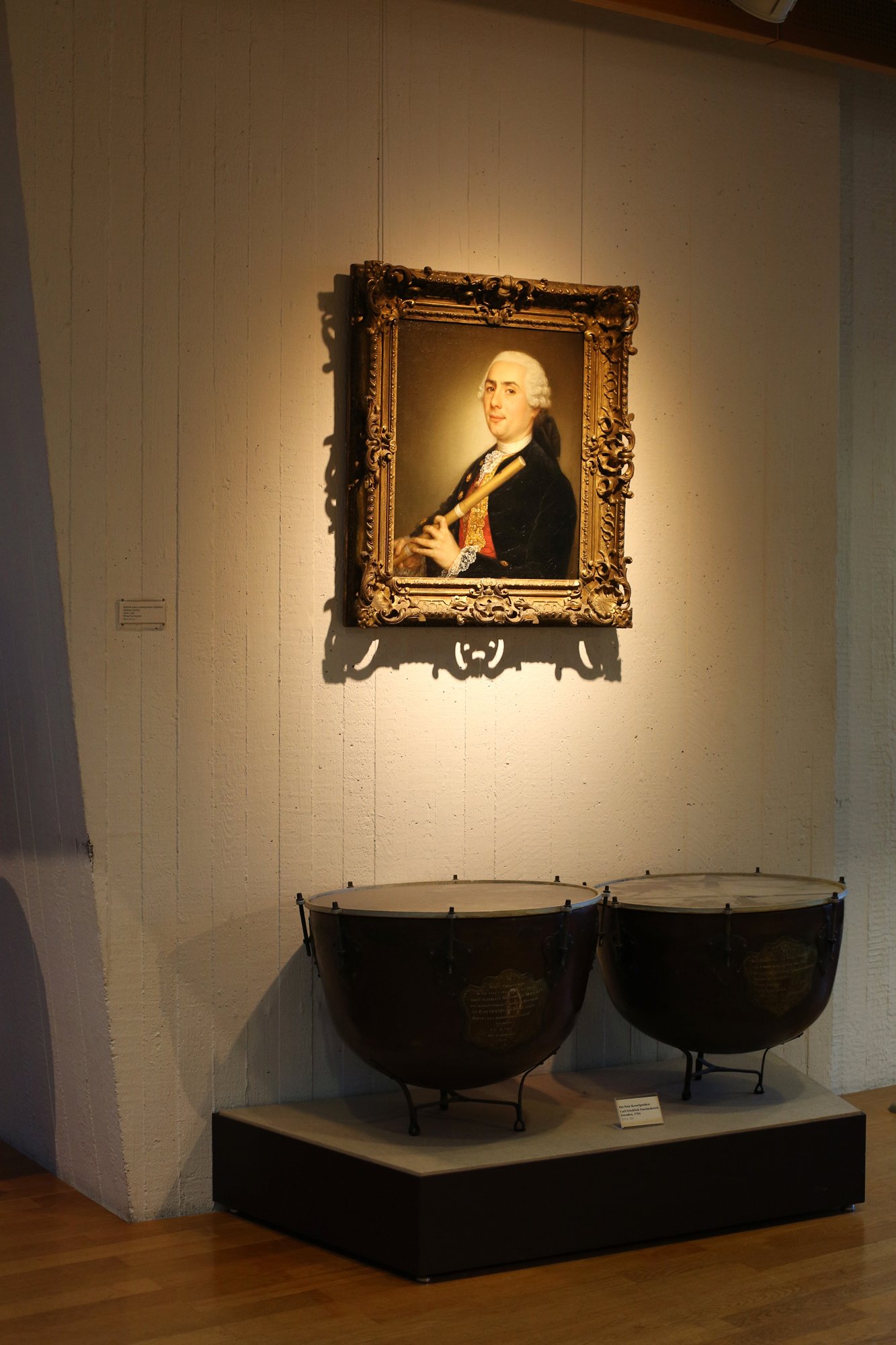 Gemälde eines Mannes aus dem 18. Jahrhundert mit Flöte an der Wand, darunter zwei historische Kesselpauken in einer Museumsvitrine.