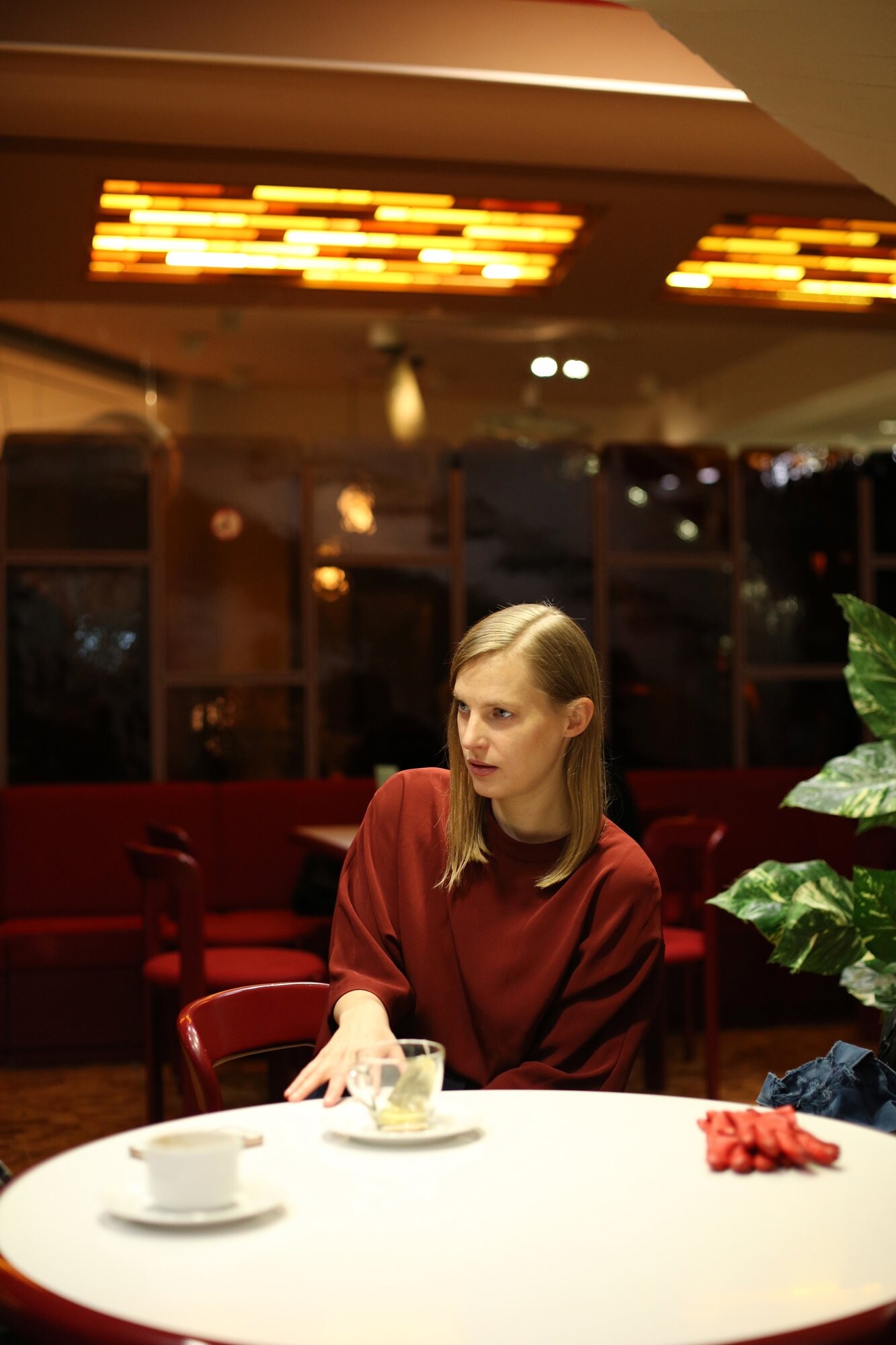 Person sitzt an einem Cafétisch mit einer Teetasse, in einem warm beleuchteten Raum mit roten Sitzbänken und Pflanzen.