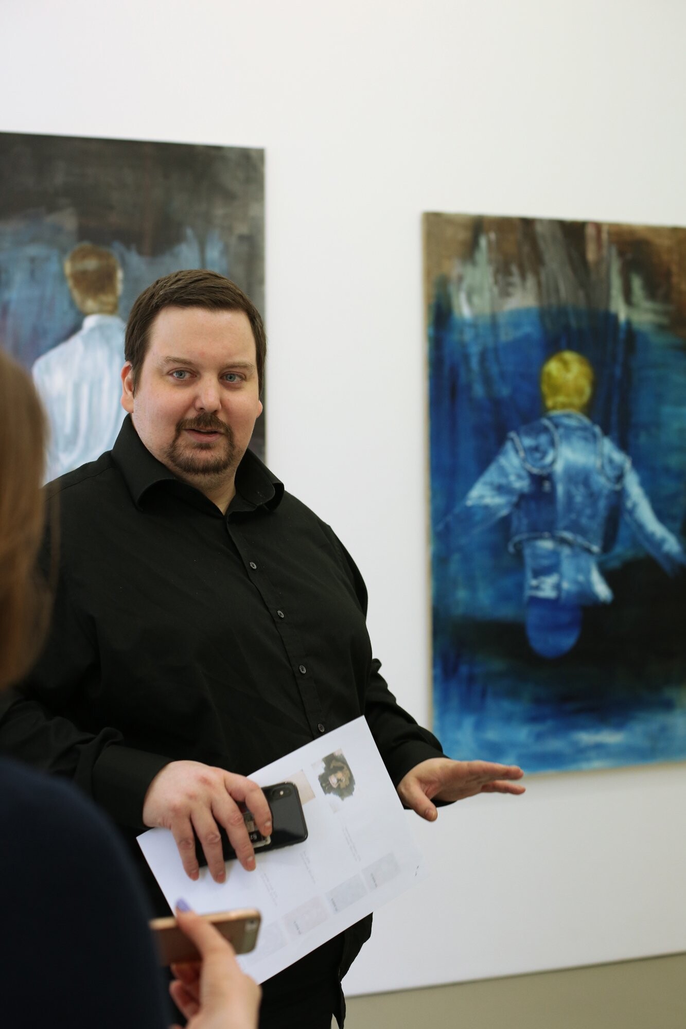Person in schwarzem Hemd spricht in einer Galerie, vor zwei großformatigen Gemälden mit Rückenfiguren in Blau- und Brauntönen.