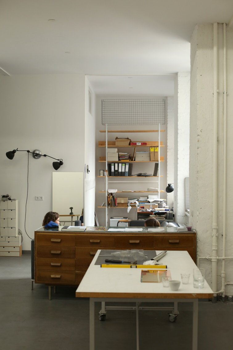 Studio space with desks, numerous documents, and a tall shelving unit at the back; two people working at their stations.