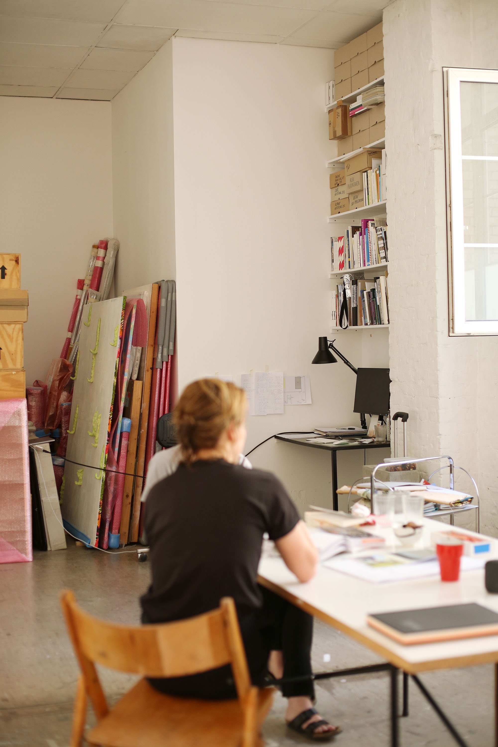 View into a studio with shelves full of books and archive boxes; two people sit at a table working, with wrapped canvases leaning in the back.