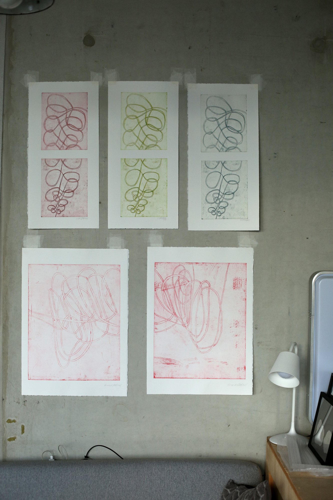 Mehrere farbige Druckgrafiken mit geschwungenen Linienmustern hängen an einer grauen Wand über einem Sofa.