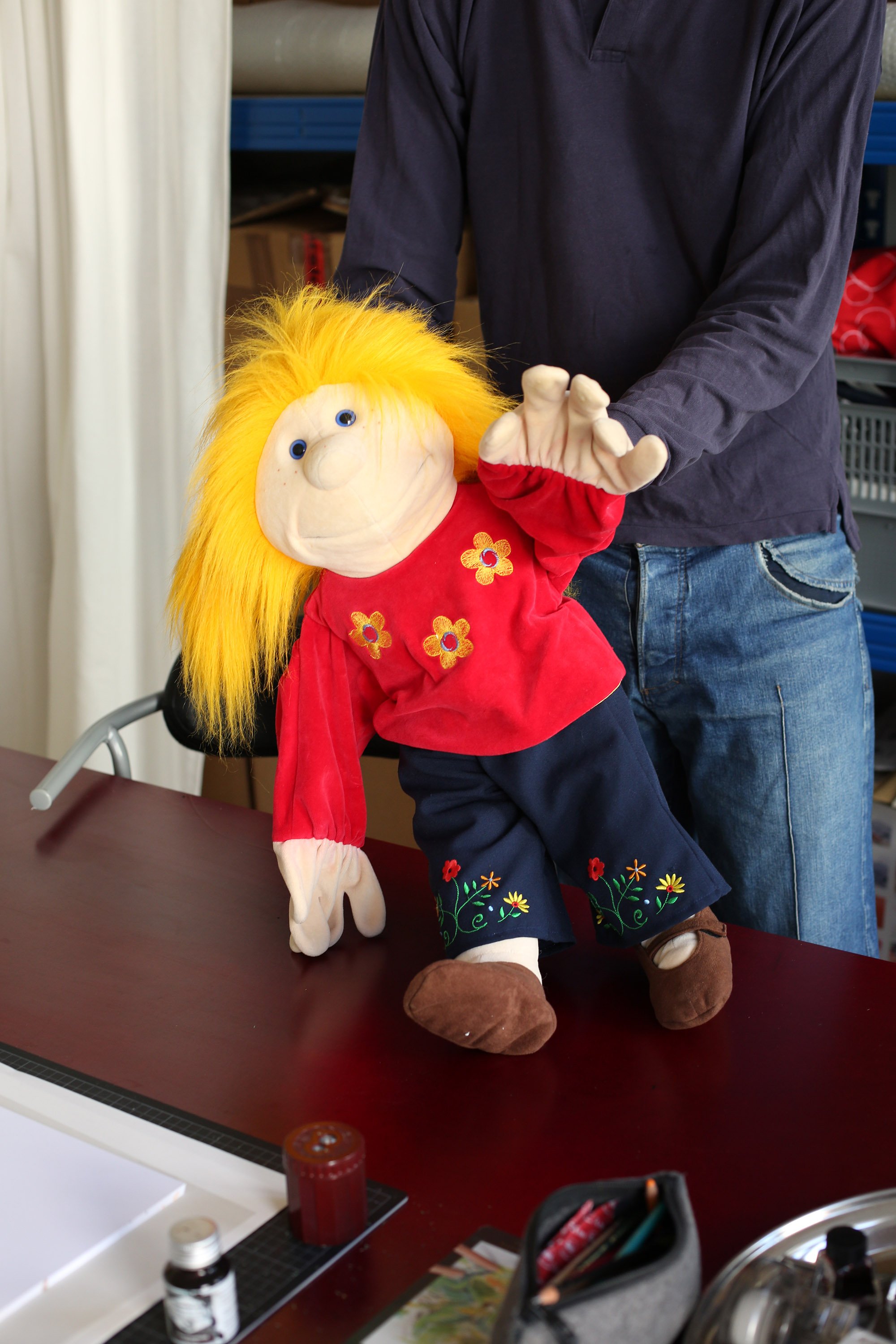 Person hält eine große Handpuppe mit gelben Haaren, rotem Hemd und dunkelblauer Hose über einen Tisch.
