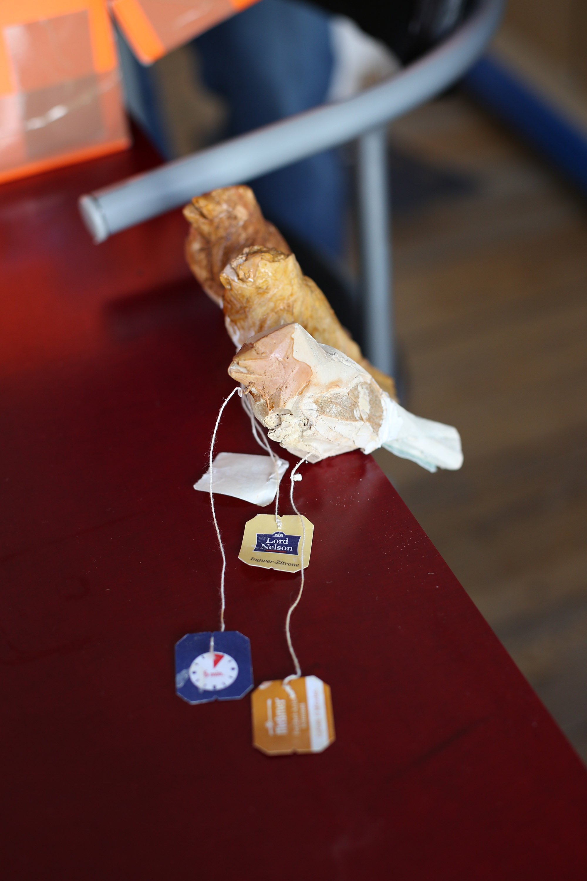 A small mixed-material sculpture rests on a table, with three tea-bag tags attached to strings.