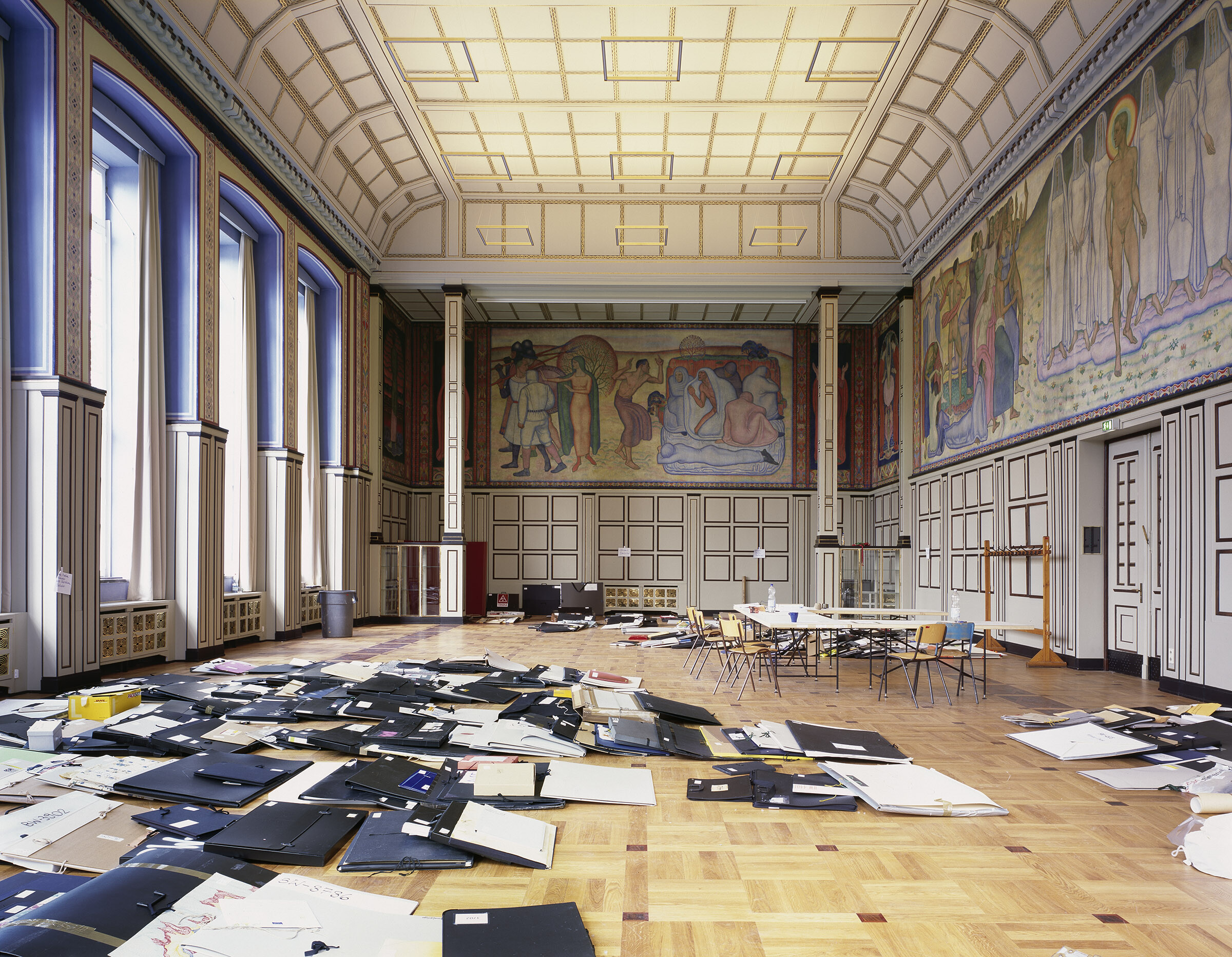 Großer, historischer Saal mit hohen Fenstern, kunstvoller Decke und Wandgemälden. Auf dem Parkettboden liegen zahlreiche Mappen und Papiere verstreut. In der Mitte stehen Tische und Stühle, als würde der Raum für Sichtungen oder Arbeitsprozesse genutzt.