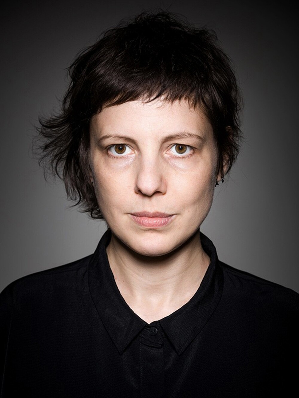 Frontal portrait of a person with short dark hair and a neutral facial expression, shown against a gray background. The person is wearing a black collared top.
