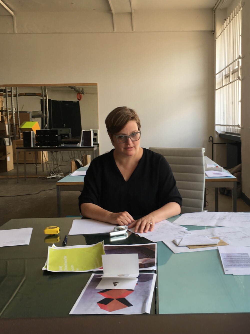 Person sitzt an einem Schreibtisch in einem Atelier oder Büro, arbeitet mit Papieren und Entwürfen.