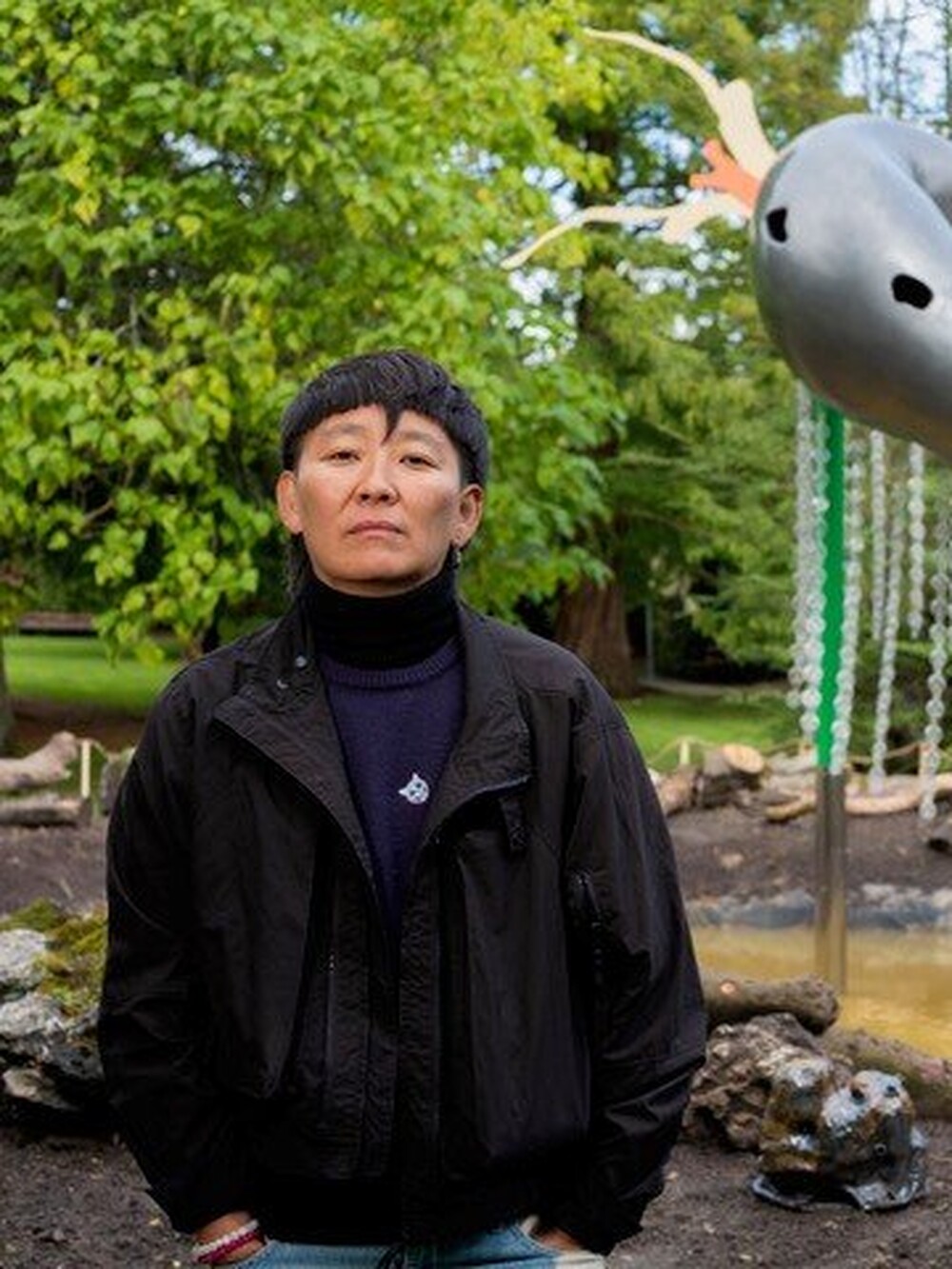 A person stands outdoors in front of a large colorful sculpture featuring hanging chain-like elements and organic shapes. Trees and a garden area can be seen in the background.