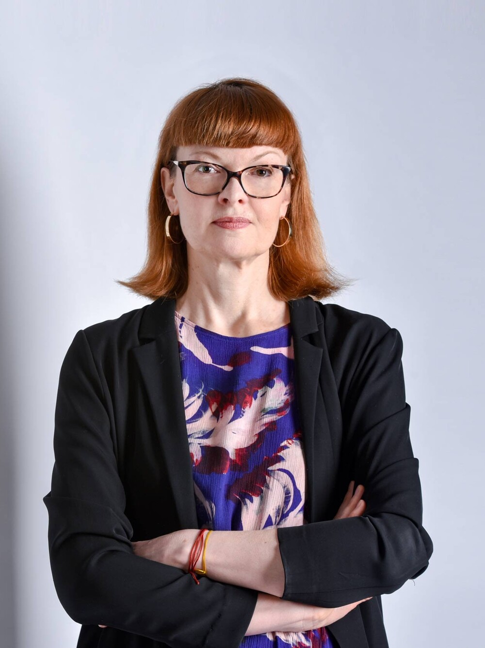 Porträt einer Person mit rötlichem, schulterlangem Haar und Brille, die mit verschränkten Armen vor einem hellen Hintergrund steht. Die Person trägt eine dunkle Jacke und ein gemustertes Oberteil.