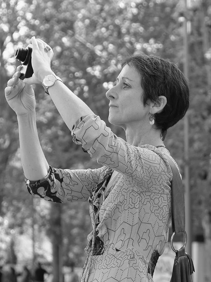 Black-and-white photograph of a woman outdoors holding a small camera or object up with both hands.