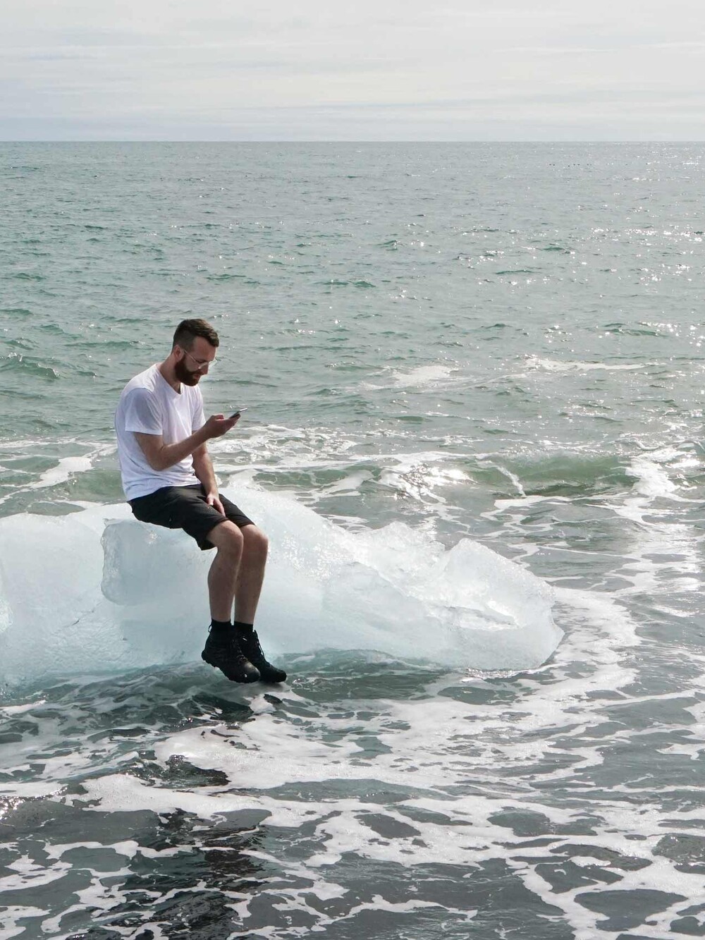 A man sits on a block of ice in the sea, looking at his smartphone as waves break around him near the shore.