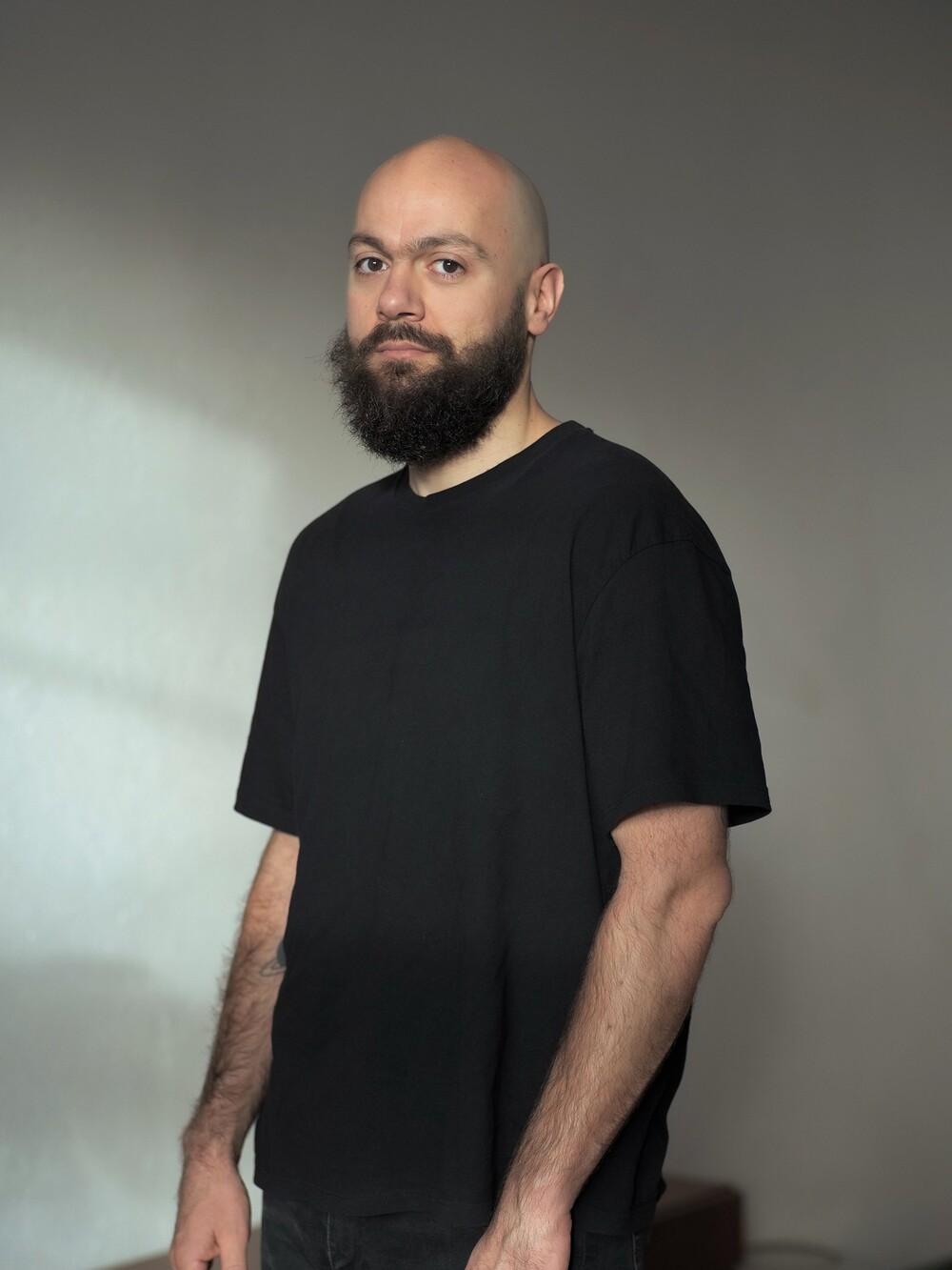 Portrait of a bald man with a full beard wearing a black T-shirt, standing in a simple indoor setting and looking directly at the camera.
