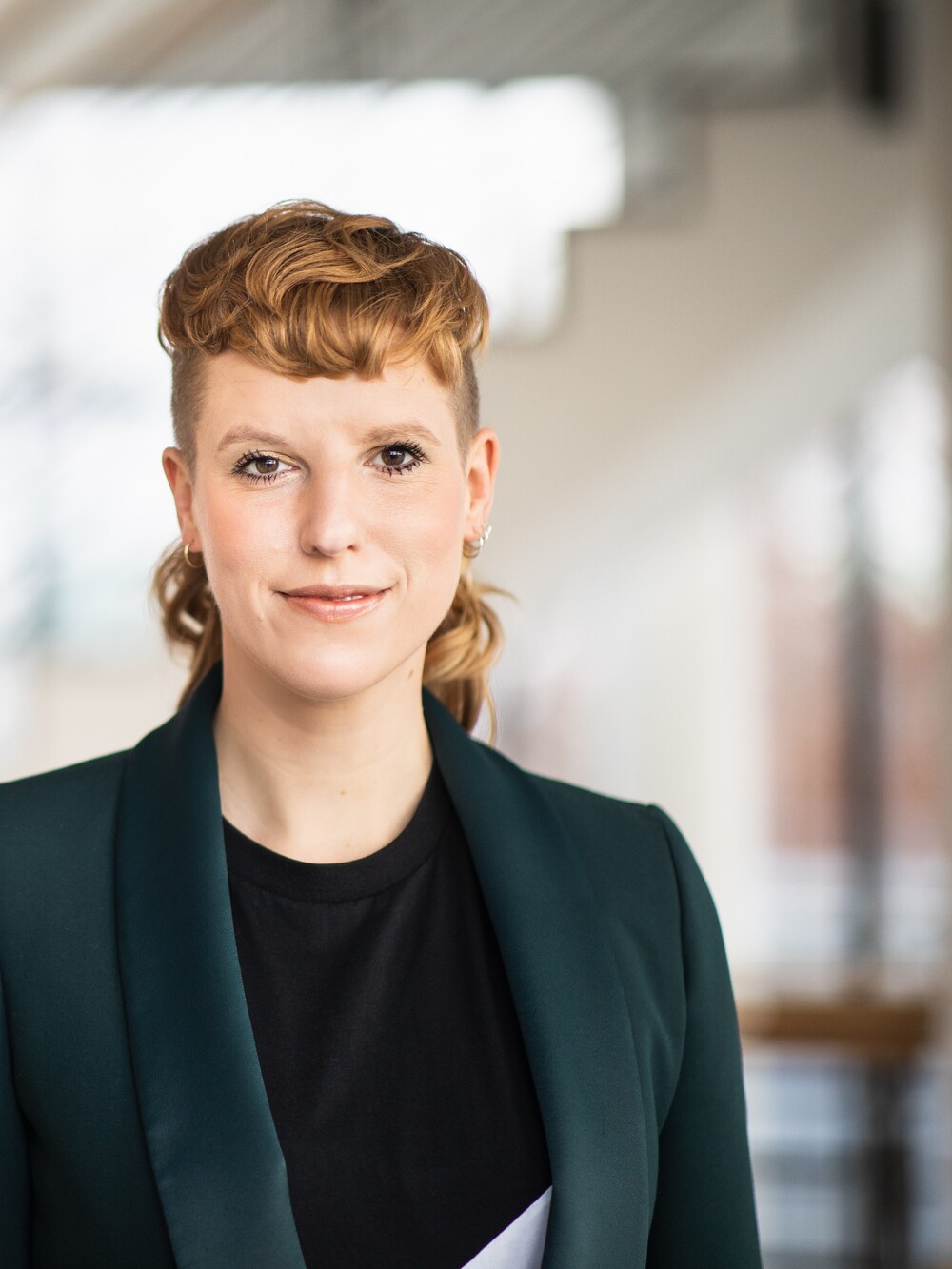 Porträt einer Frau mit hochgesteckten, kurzen Haaren und grünem Blazer vor einem hellen, unscharfen Innenraum.