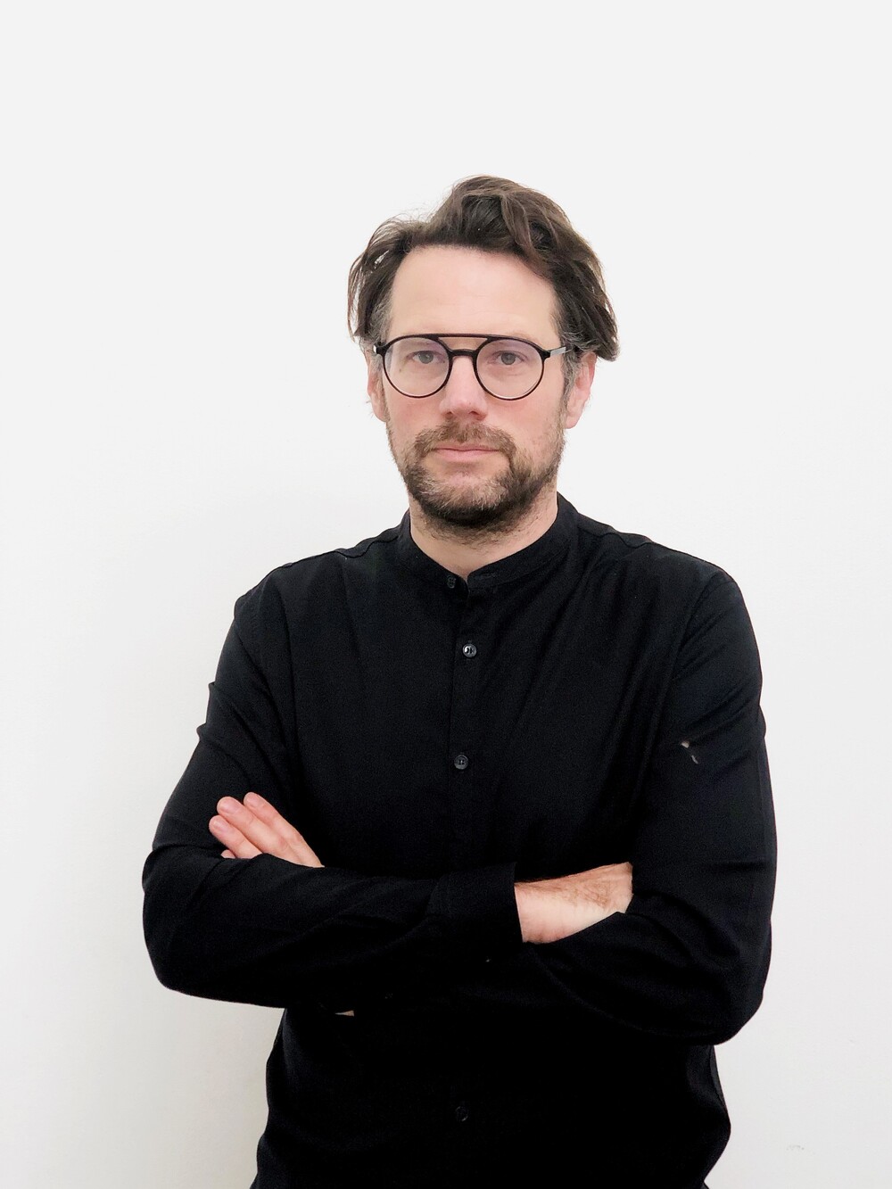 Portrait of a man with glasses and a beard, standing with crossed arms in front of a white background.