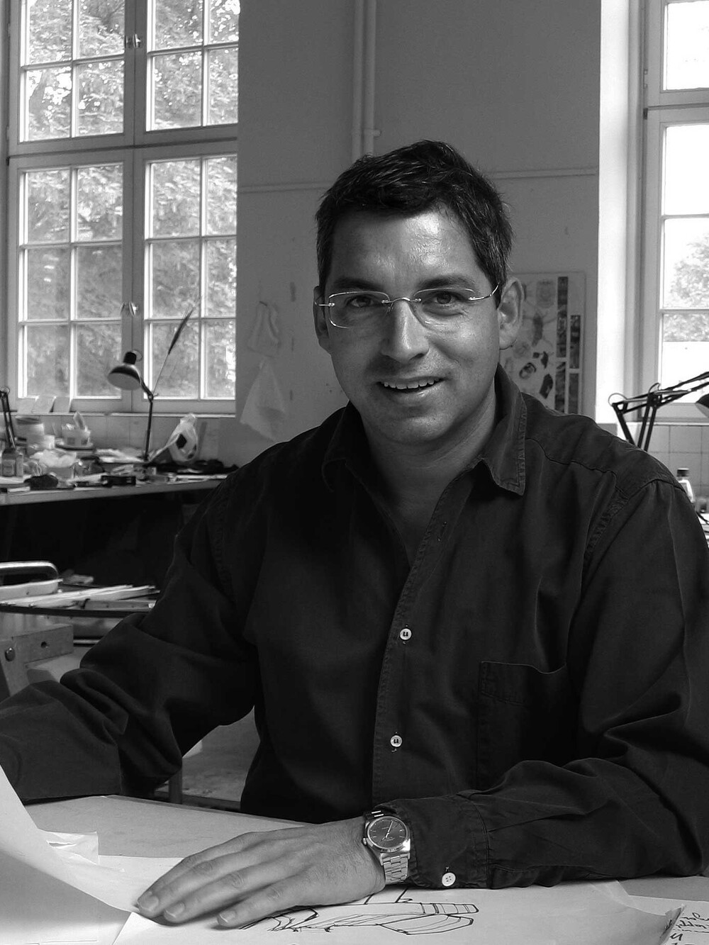 Schwarzweißporträt eines Mannes mit Brille, der an einem Arbeitstisch sitzt und Zeichnungen in einem Atelier betrachtet.