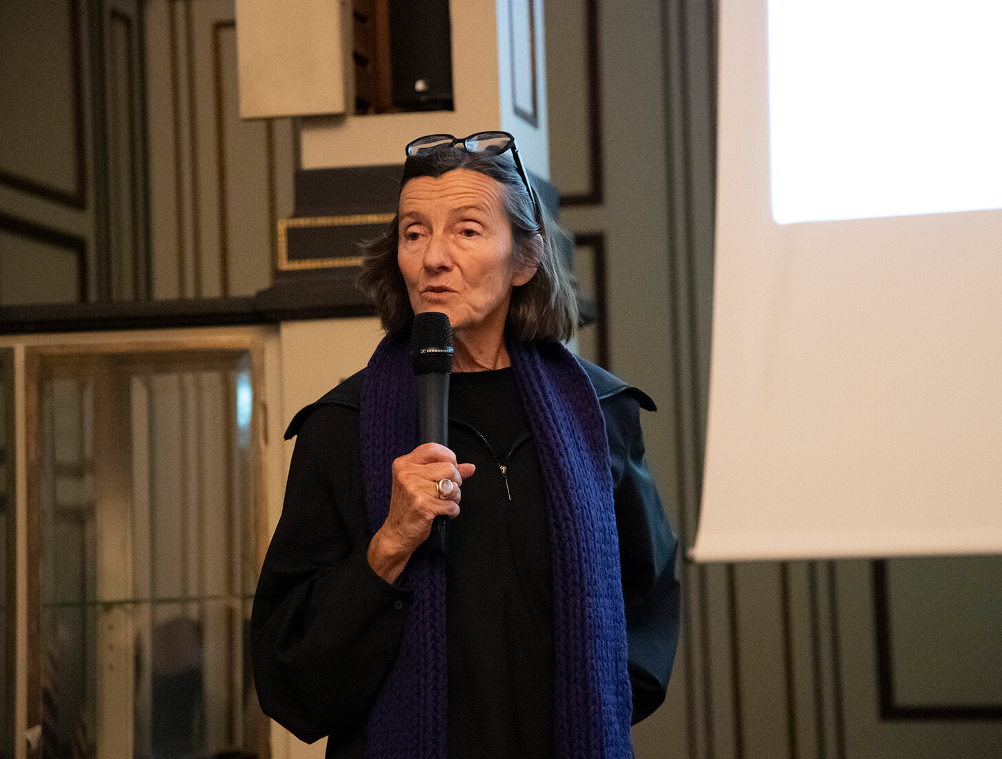 A woman stands in a historic interior and speaks into a handheld microphone. She is wearing dark clothing and a purple scarf, with glasses resting on her head. A projection screen is visible beside her. The room features decorative wall paneling and appears to be a venue for a lecture or talk.