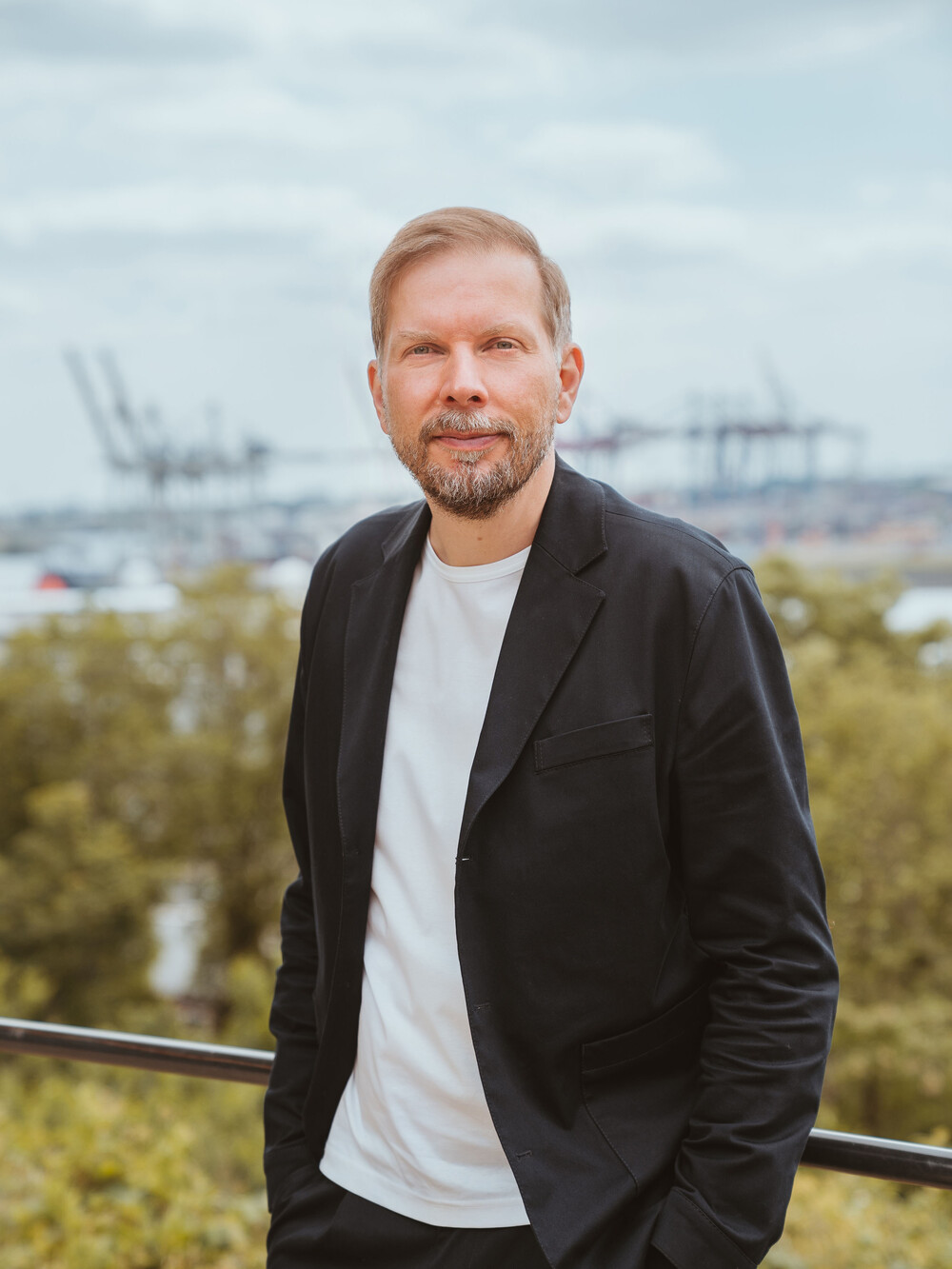 A man with short blond hair and a beard stands outdoors on a terrace. He is wearing a black blazer over a white T-shirt with his hands in his pockets. In the background there are trees, water, and harbor cranes.