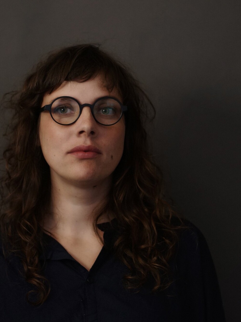 Porträt einer Frau mit lockigem Haar und runder Brille vor dunklem, unscharfem Hintergrund.