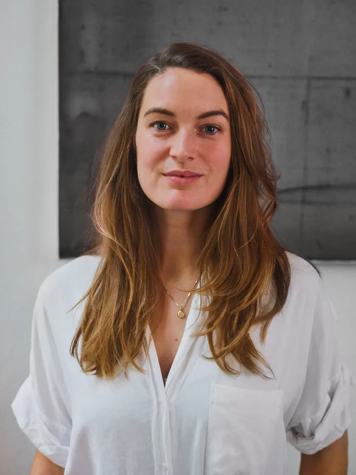 Portrait of a woman with long, loose hair wearing a white blouse, standing in front of a gray wall.